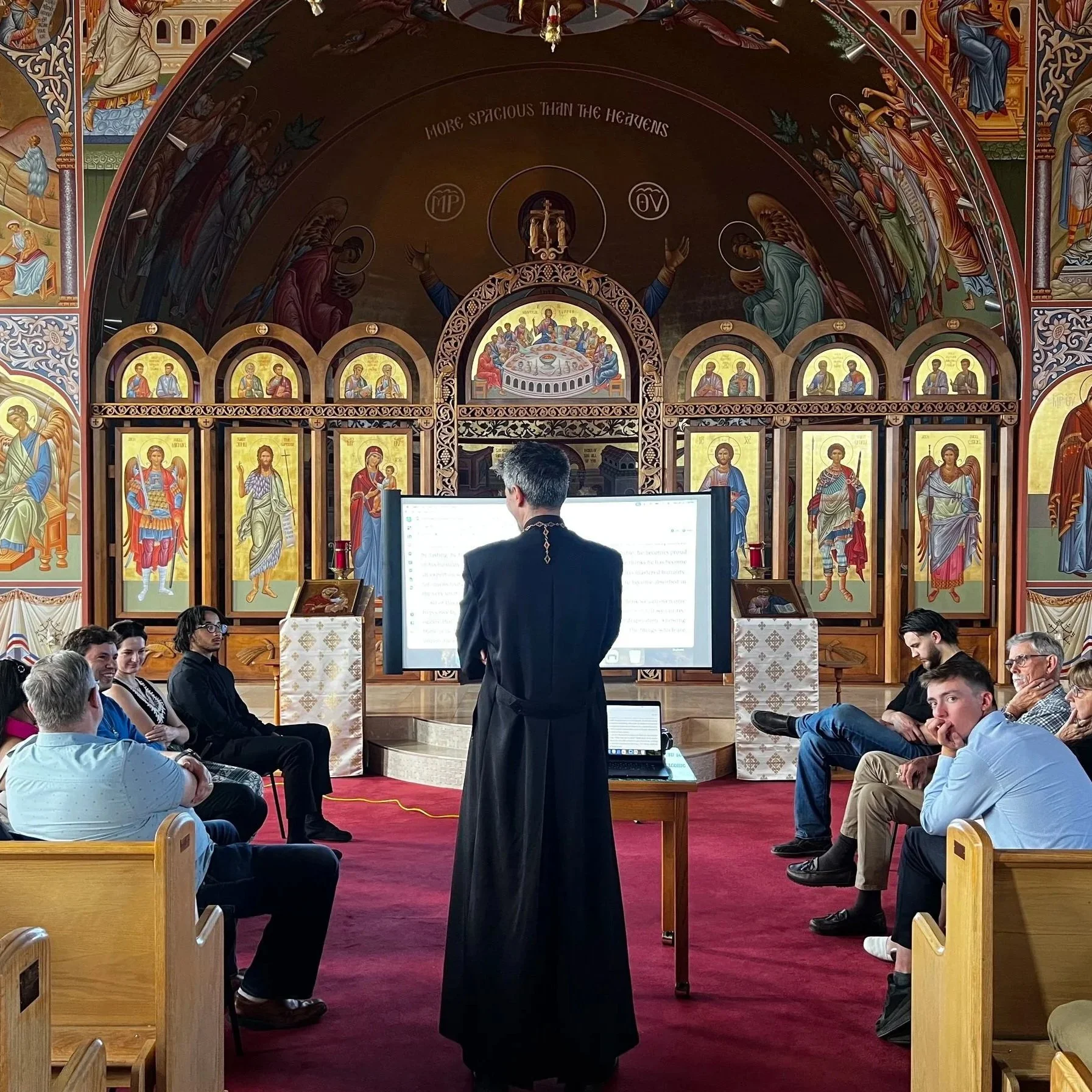 A religious service in a church with people seated and a priest in black robes standing at the altar. The church interior features ornate woodwork and religious icons, including an iconostasis with images of saints. The congregation is seated on wooden pews, and a computer monitor is visible at the front.