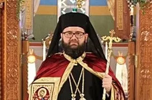 The Right Reverend Andrei (Hoarste) PhD Bishop of Cleveland and Vicar Bishop of the Romanian Orthodox Episcop dressed in religious or ceremonial robes with a head covering and glasses, holding a staff, standing indoors with ornate wooden furnishings.