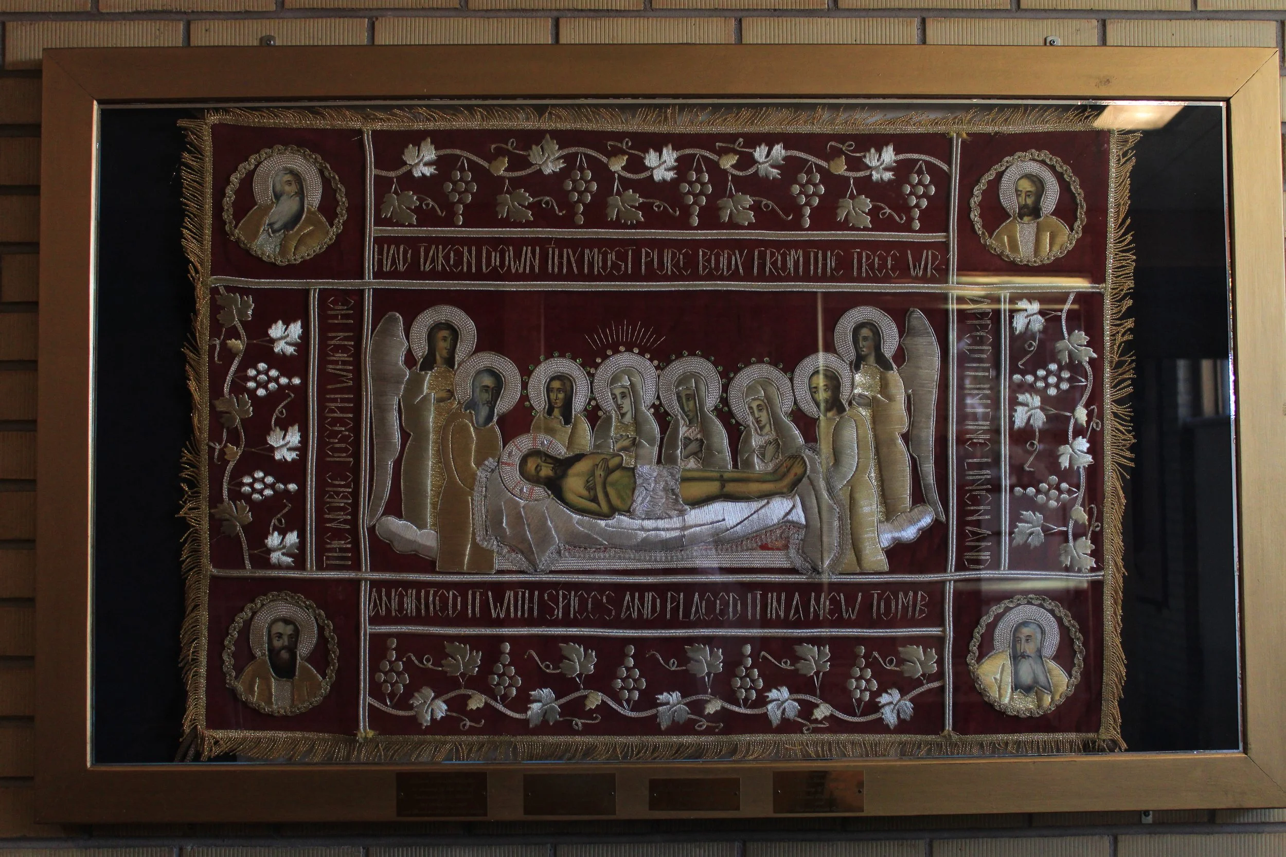 Religious embroidery depicting the body of Jesus Christ laid in a tomb surrounded by saints and angels, with inscriptions and decorative elements.