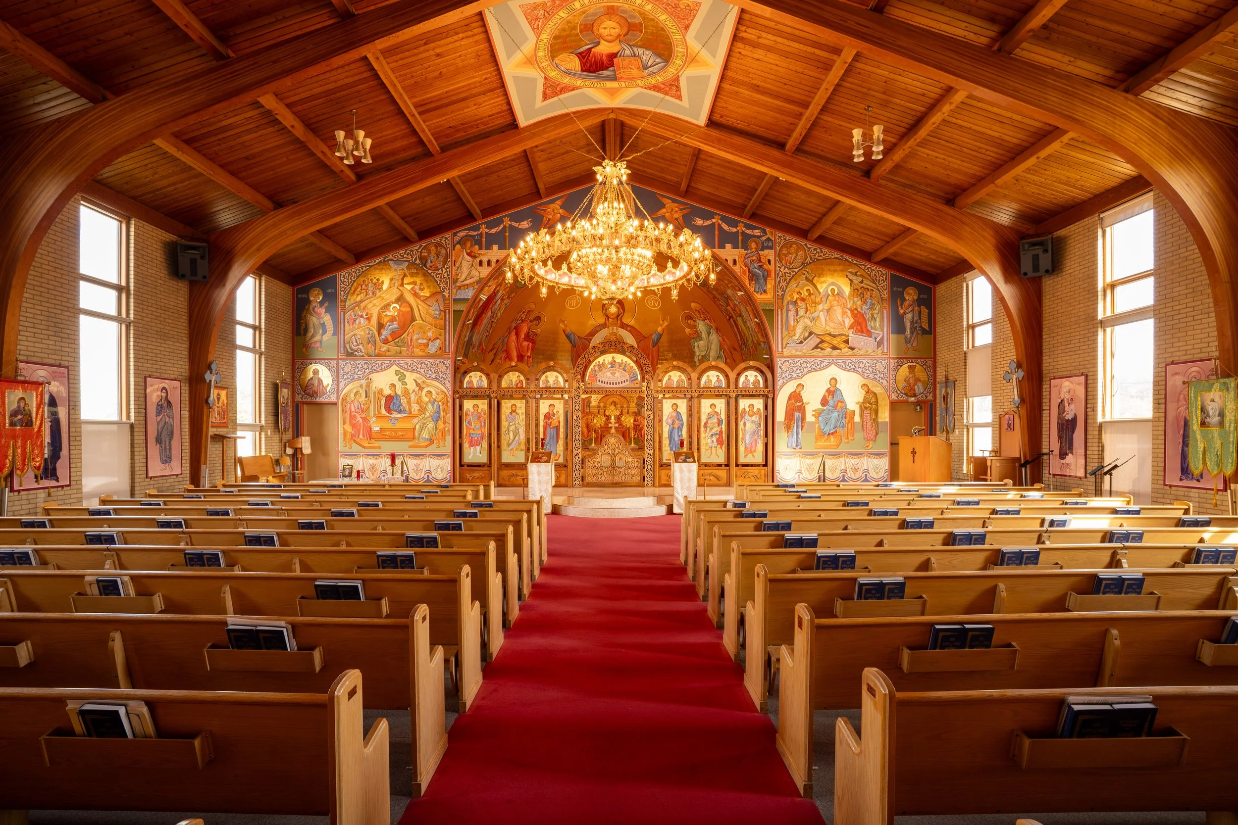 Interior of a Christian church with wooden vaulted ceiling, chandeliers, stained glass windows, religious icons, and wooden pews.