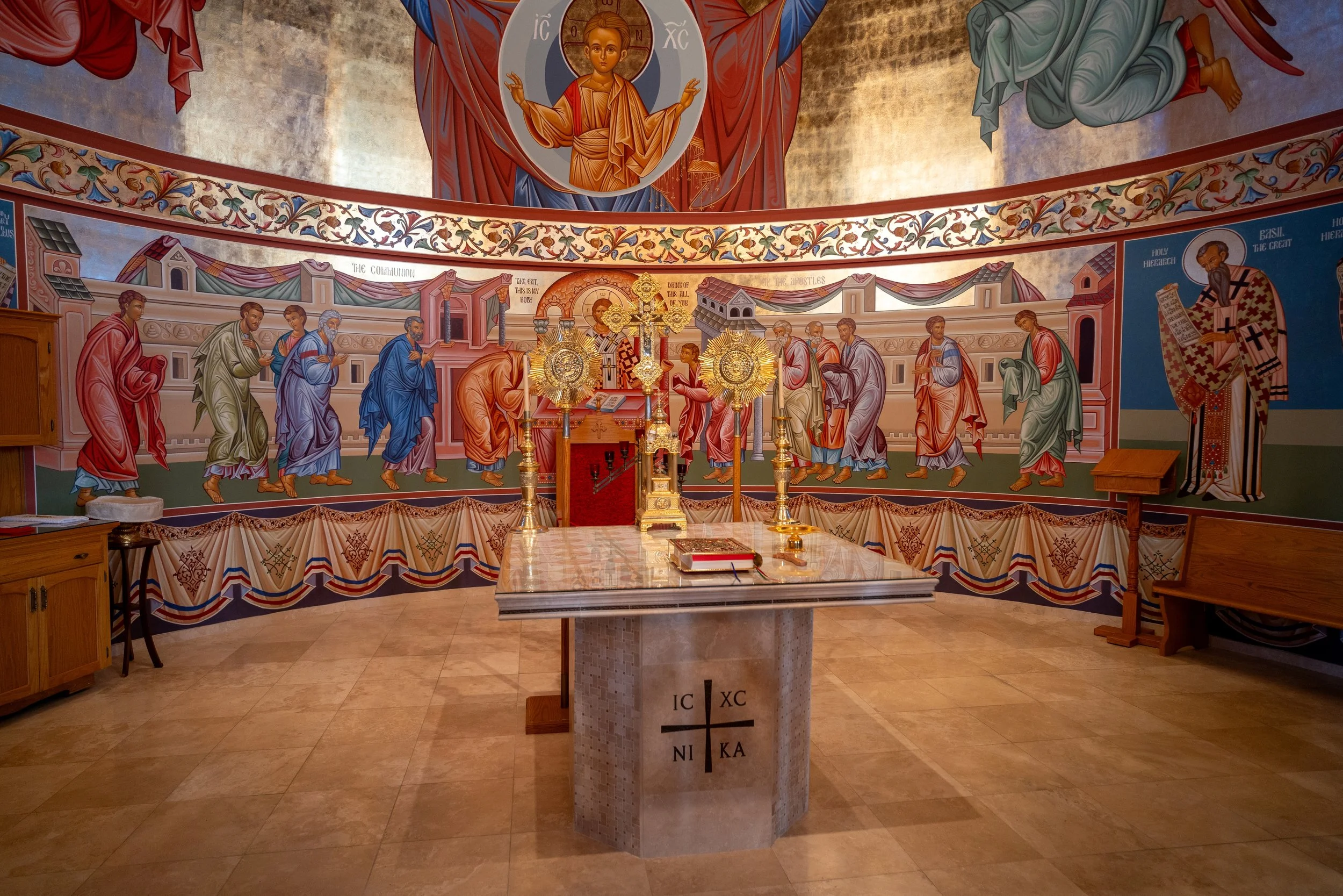 Interior of an Orthodox Christian church with religious iconography, including a central altar with gold artifacts, religious paintings on the walls and ceiling, and a prominent depiction of Jesus Christ in the domed ceiling.