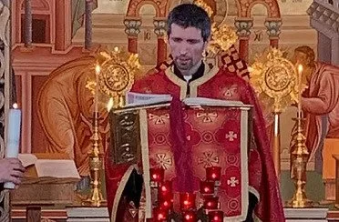 Parish priest, Father Emmanuel Cristian Frunzulica, wearing a red robe and standing at an ornate altar during a religious ceremony, with candles and religious icons in the background.