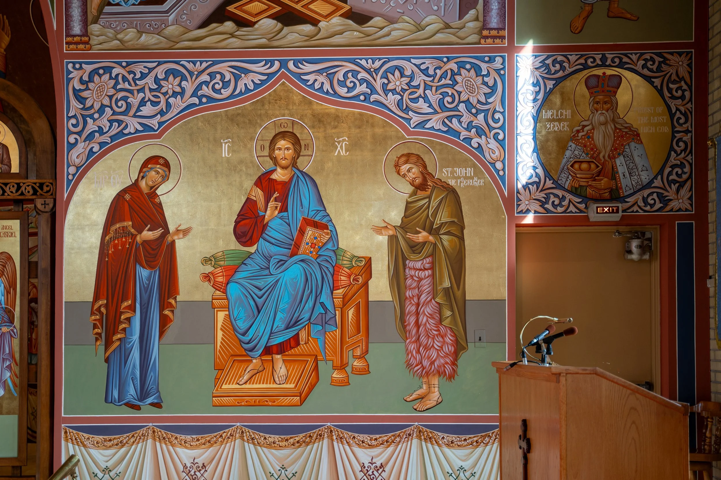 An Orthodox Christian religious mural depicting Jesus Christ in the center, with the Virgin Mary on the left and St. John the Forerunner on the right. The mural is decorated with intricate floral and geometric patterns in vibrant colors.