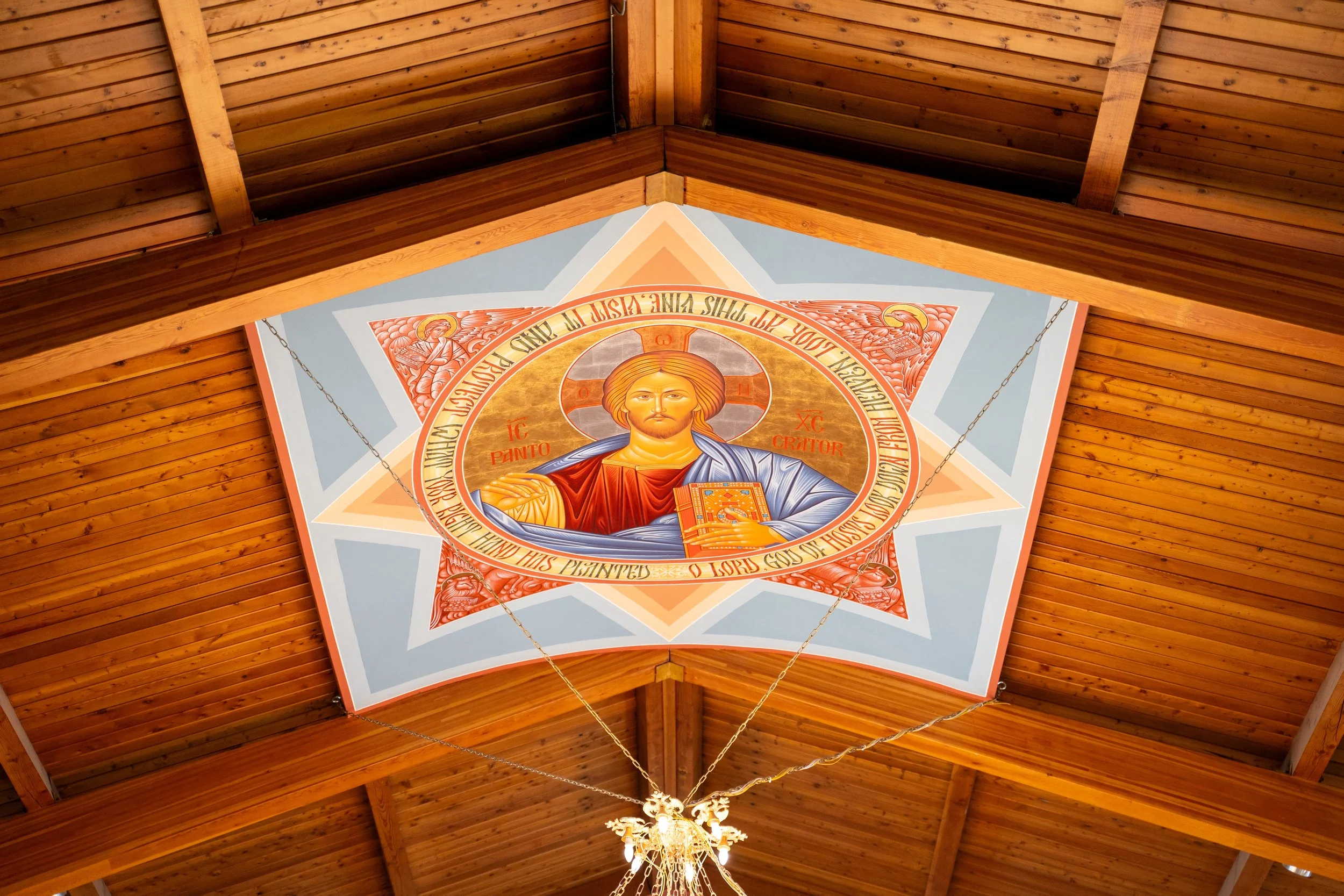 Christian religious artwork on the ceiling in a church. The artwork features Jesus Christ with a halo, holding a book, with Greek symbols and religious text surrounding him. The ceiling is made of wooden panels, and there is a chandelier hanging below.