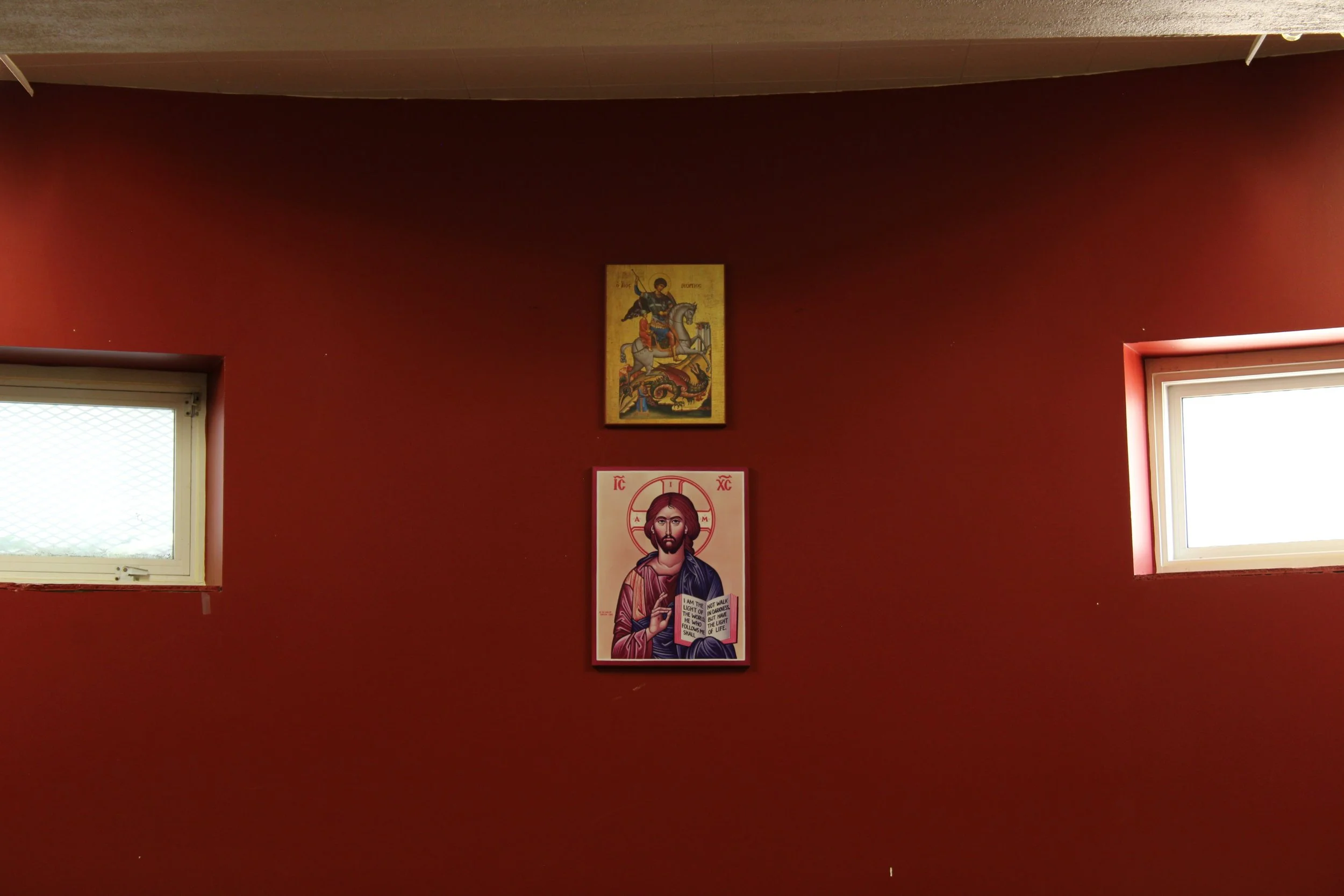 Interior view of a room with red walls, two small windows on either side, and religious icons hanging in the center, including an icon of Jesus Christ and a depiction of a saint on a horse.