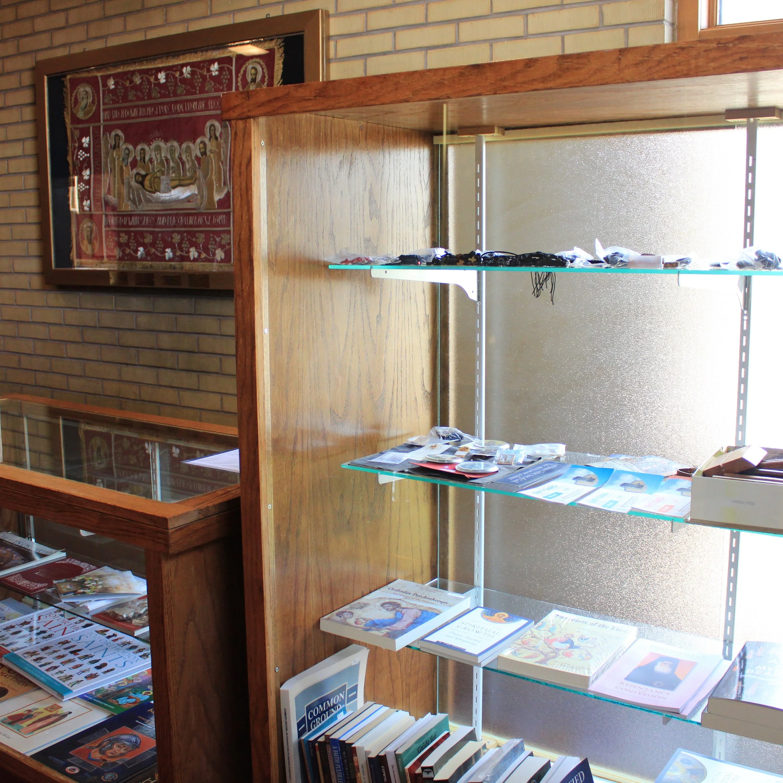 Wooden display shelf with religious artwork, books, and pamphlets against a brick wall and a window.