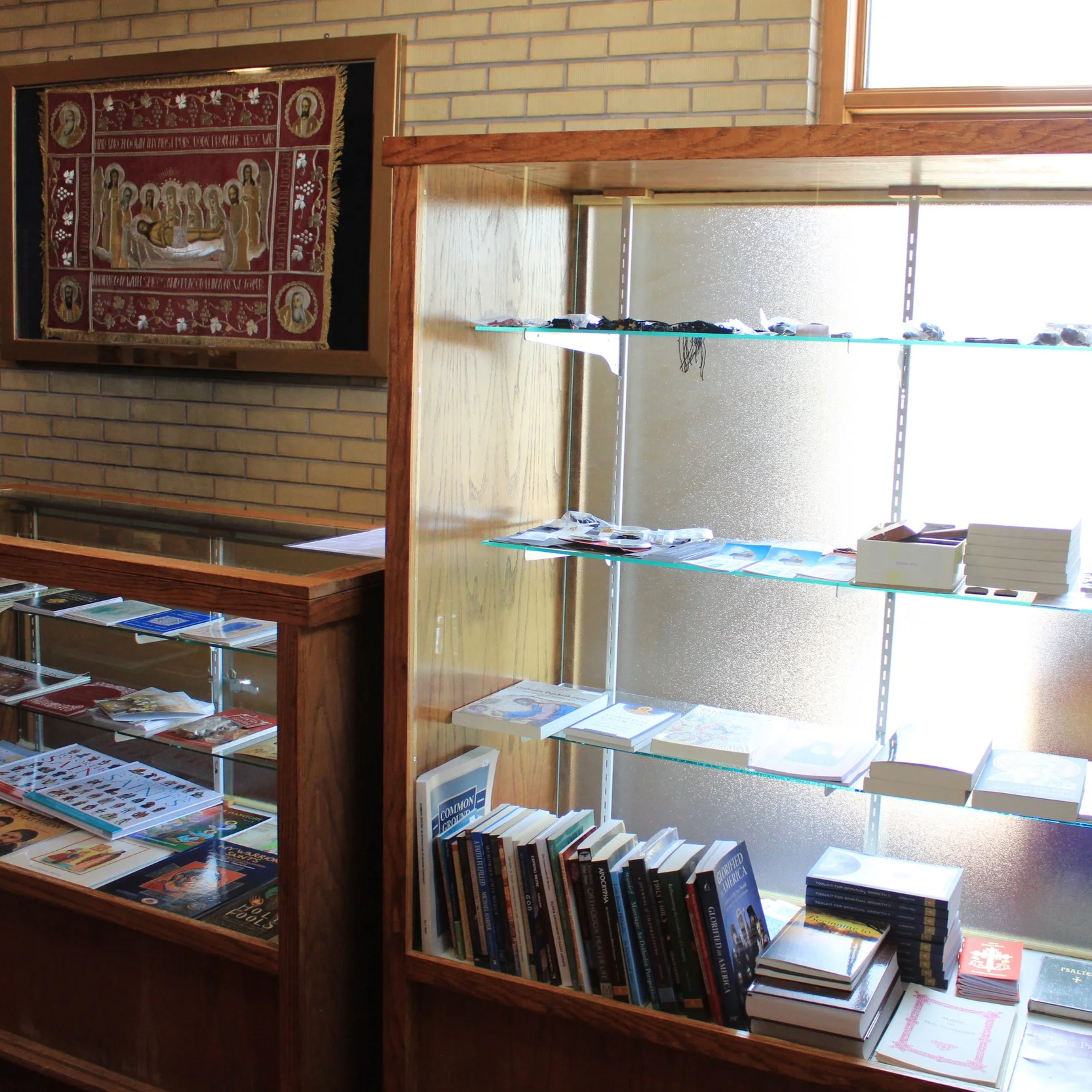 Interior of a bookstore with display shelves filled with books, and a religious embroidery hanging on a brick wall.
