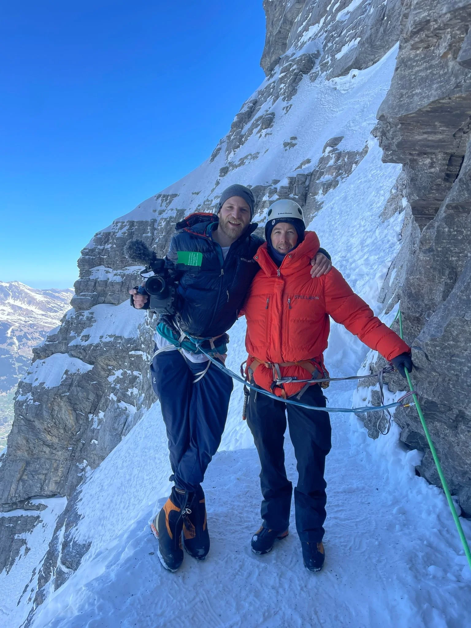 Two climbers in mountain gear standing on a snowy ledge next to a rock face with a mountain landscape in the background.