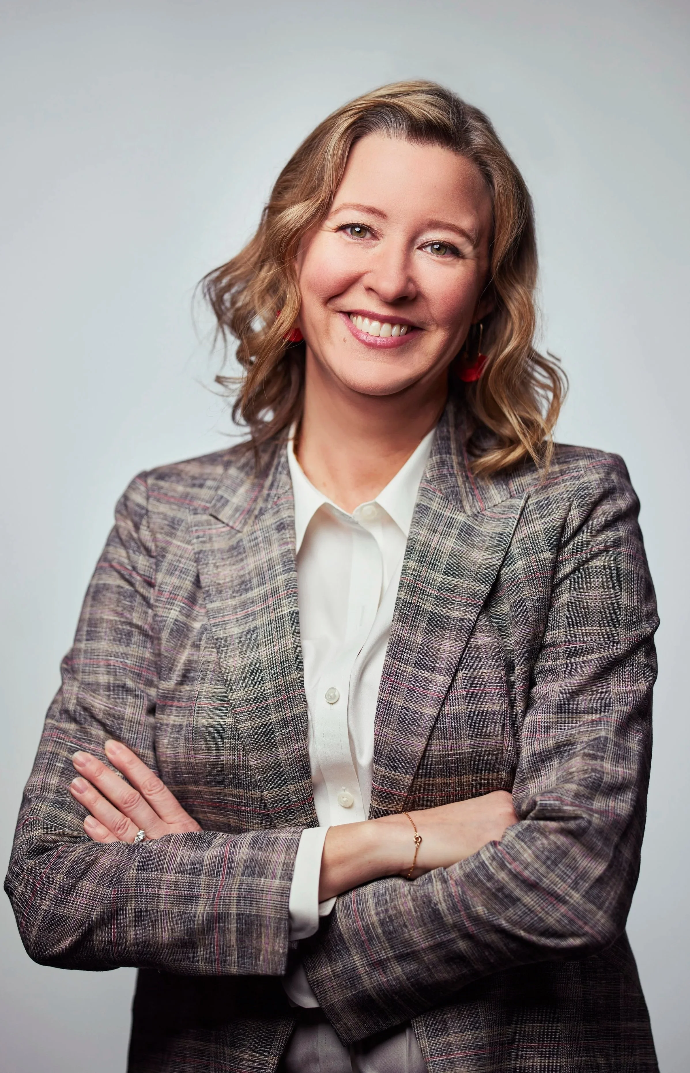 A professional woman with shoulder-length wavy hair, wearing a plaid blazer over a white shirt, stands with arms crossed and smiling at the camera against a neutral background.