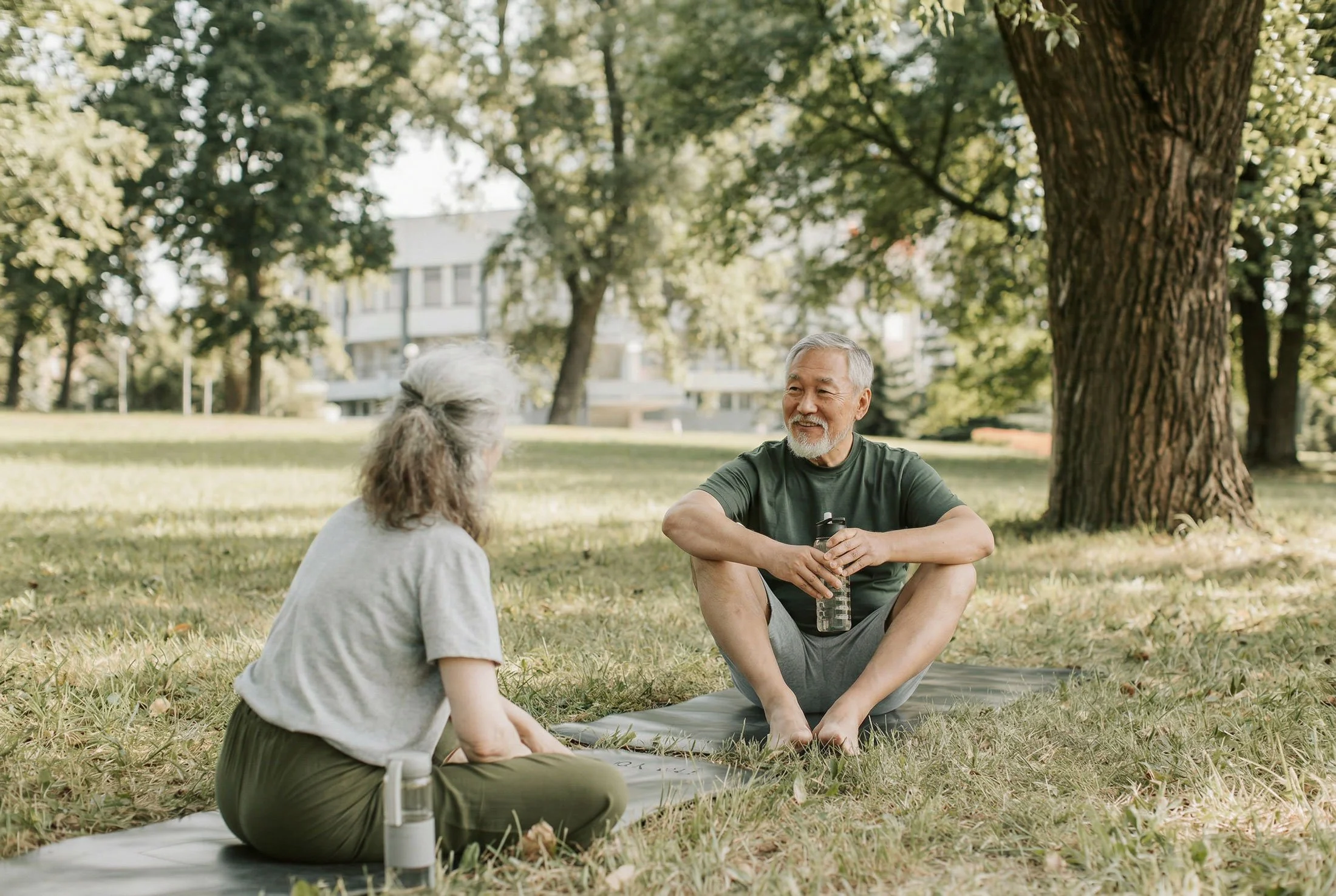 Two midlife adults sitting on yoga mats in a sunny park, sharing a relaxed conversation under shaded trees