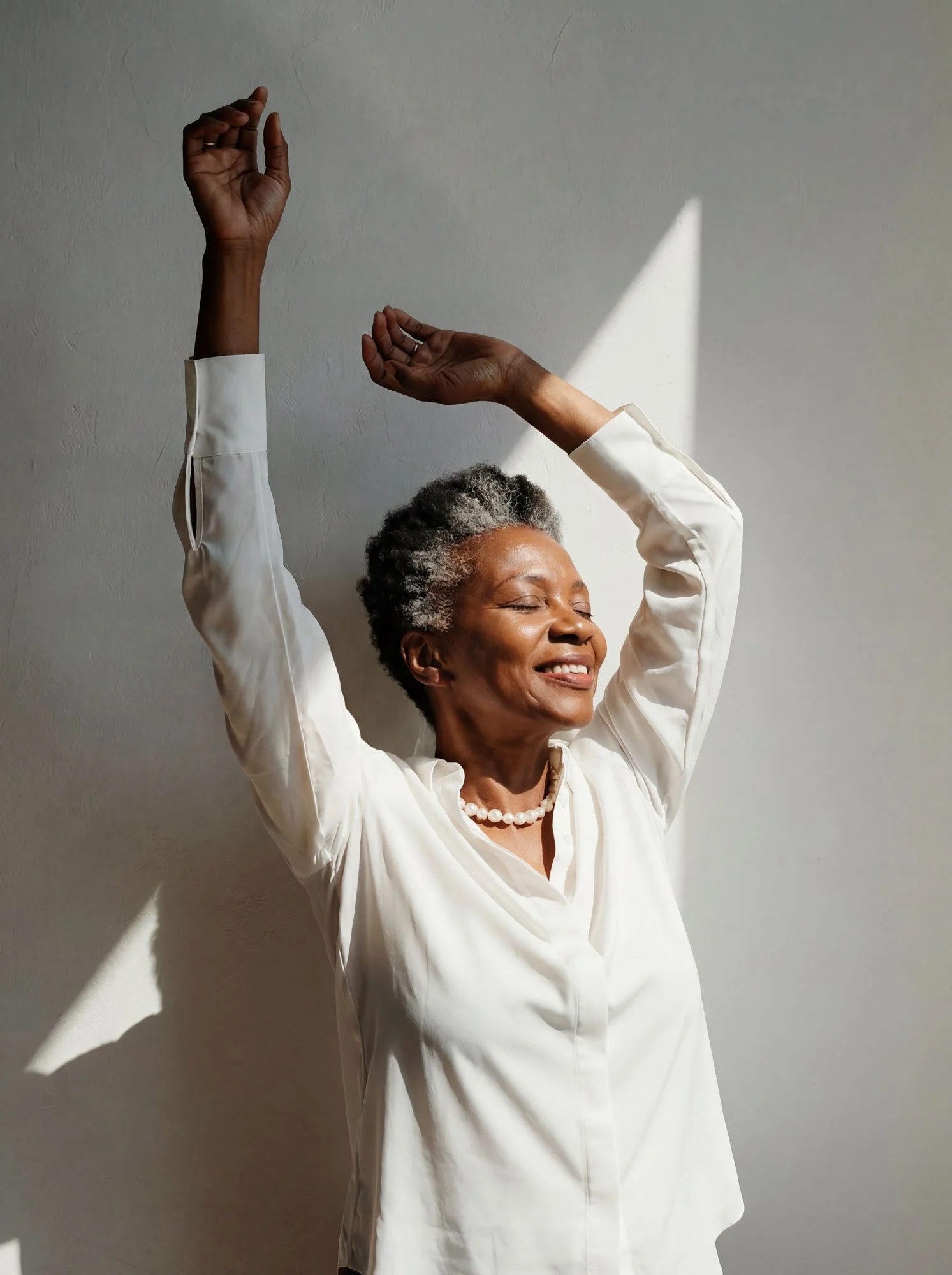 Woman in her 60s with natural grey hair, eyes closed, smiling joyfully with arms raised in a bright minimal space