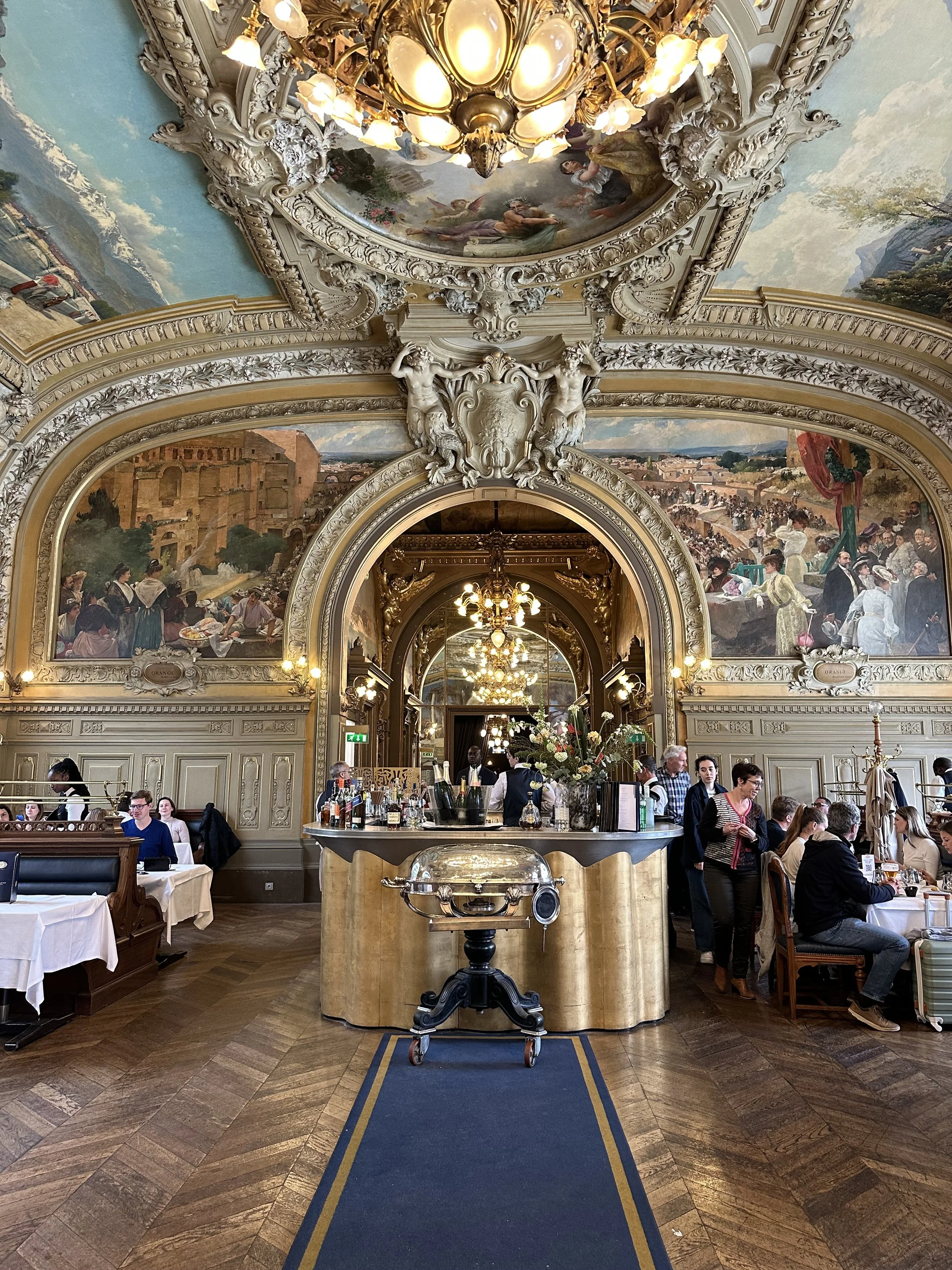The ornate interior of a historic restaurant with detailed ceiling murals, chandeliers, and a bar area in the center. People are seated at tables, enjoying meals, with waitstaff attending to guests.