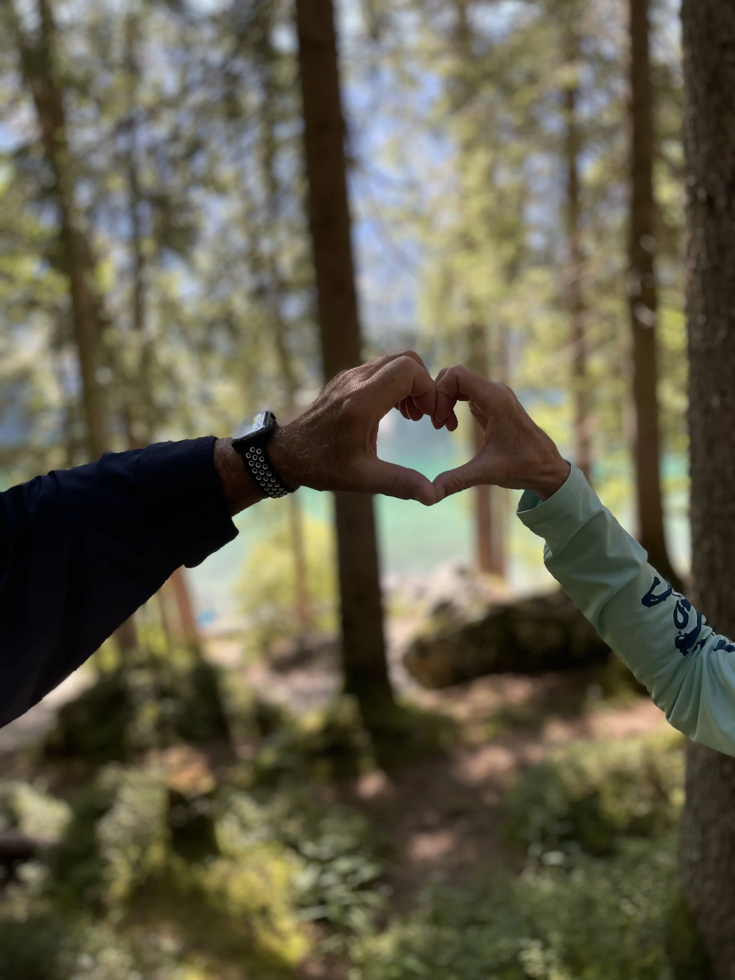 Two people in outdoor clothing form a heart shape with their hands in front of a forest scene with tall trees and sunlight filtering through the leaves.