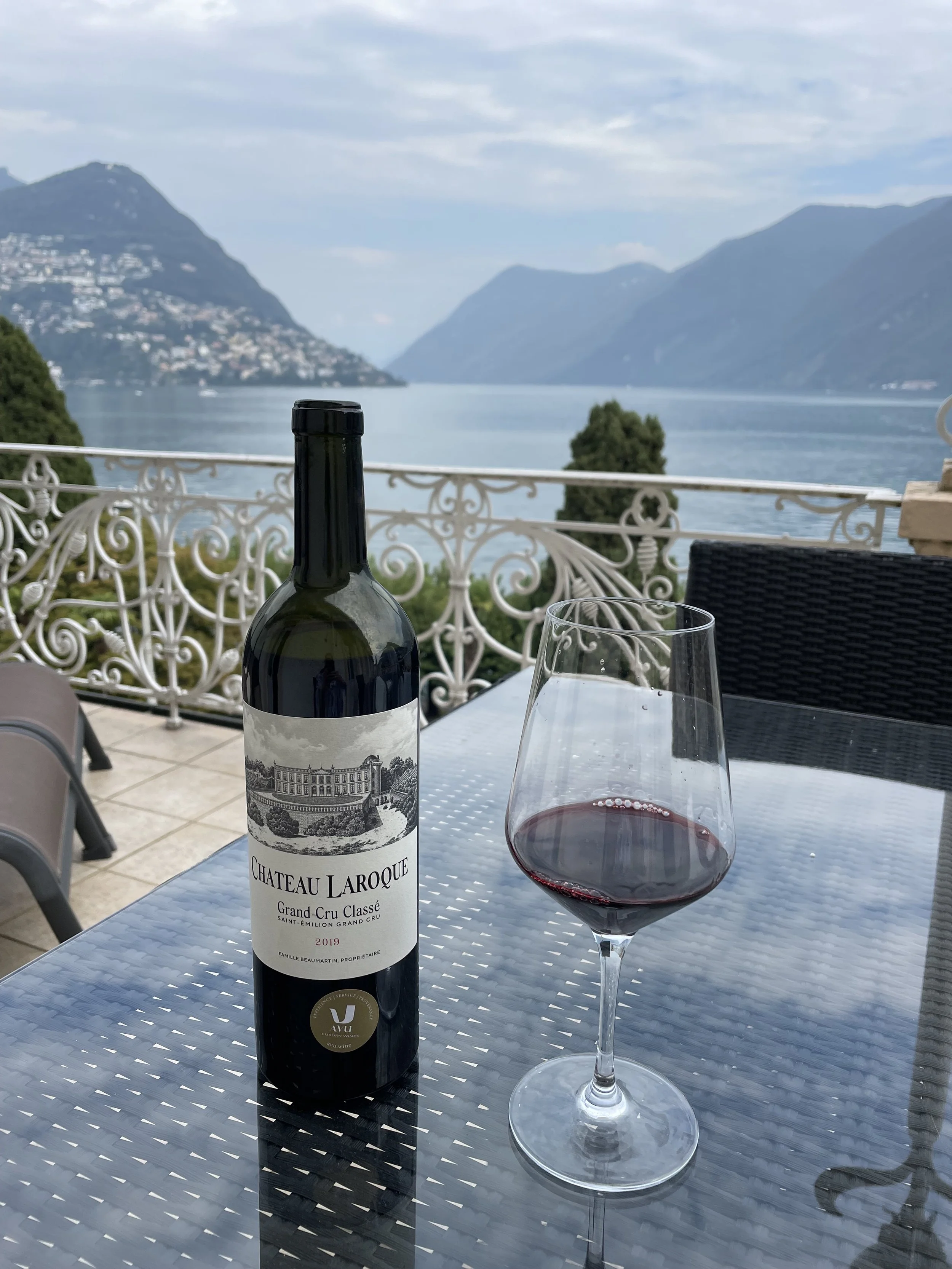 A bottle of Château Laroque red wine and a glass of red wine on a table overlooking a lake and mountain landscape.