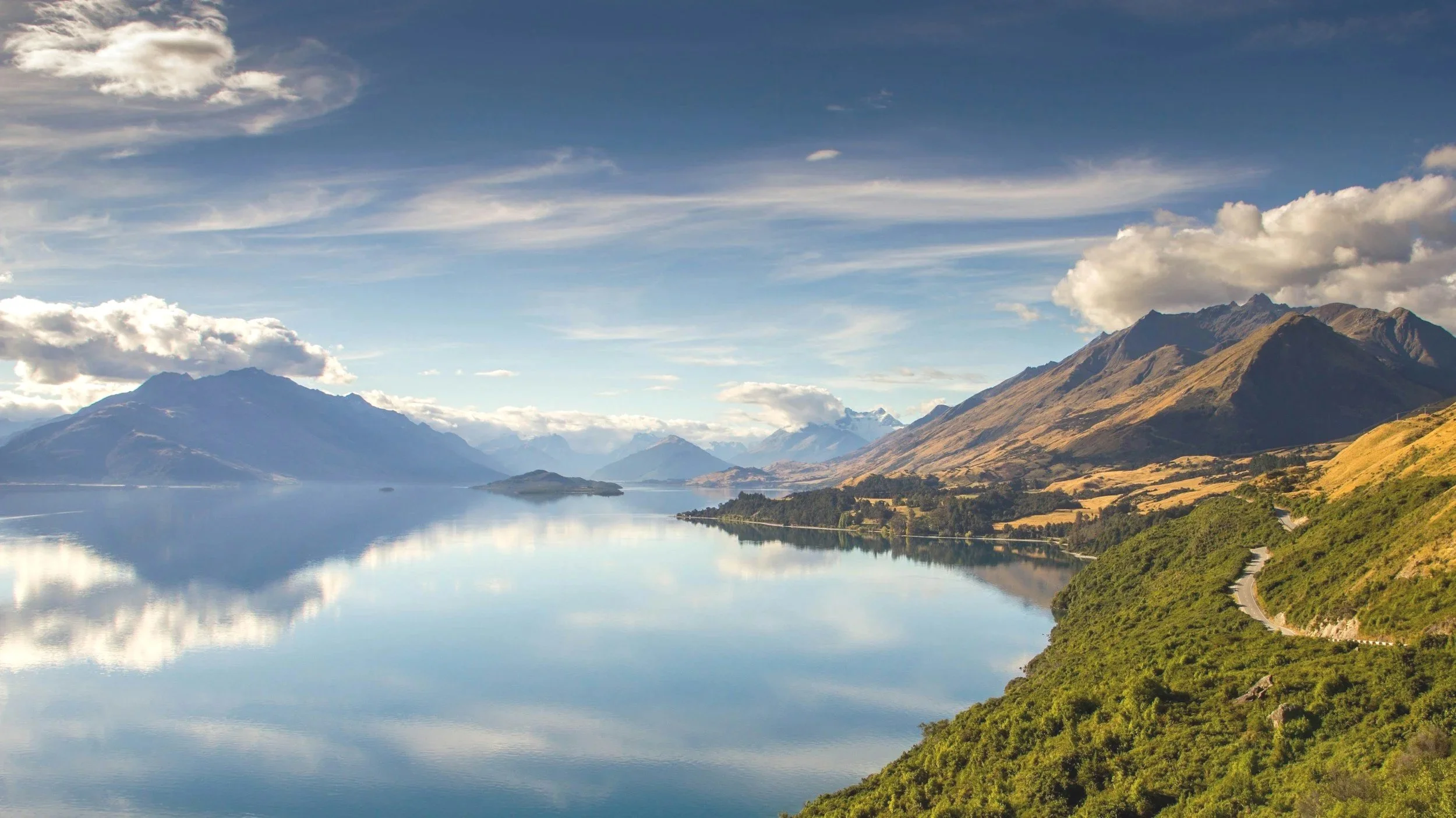 mountains and a lake with still water reflecting the sky and clouds, surrounded by green rolling hills and a winding road