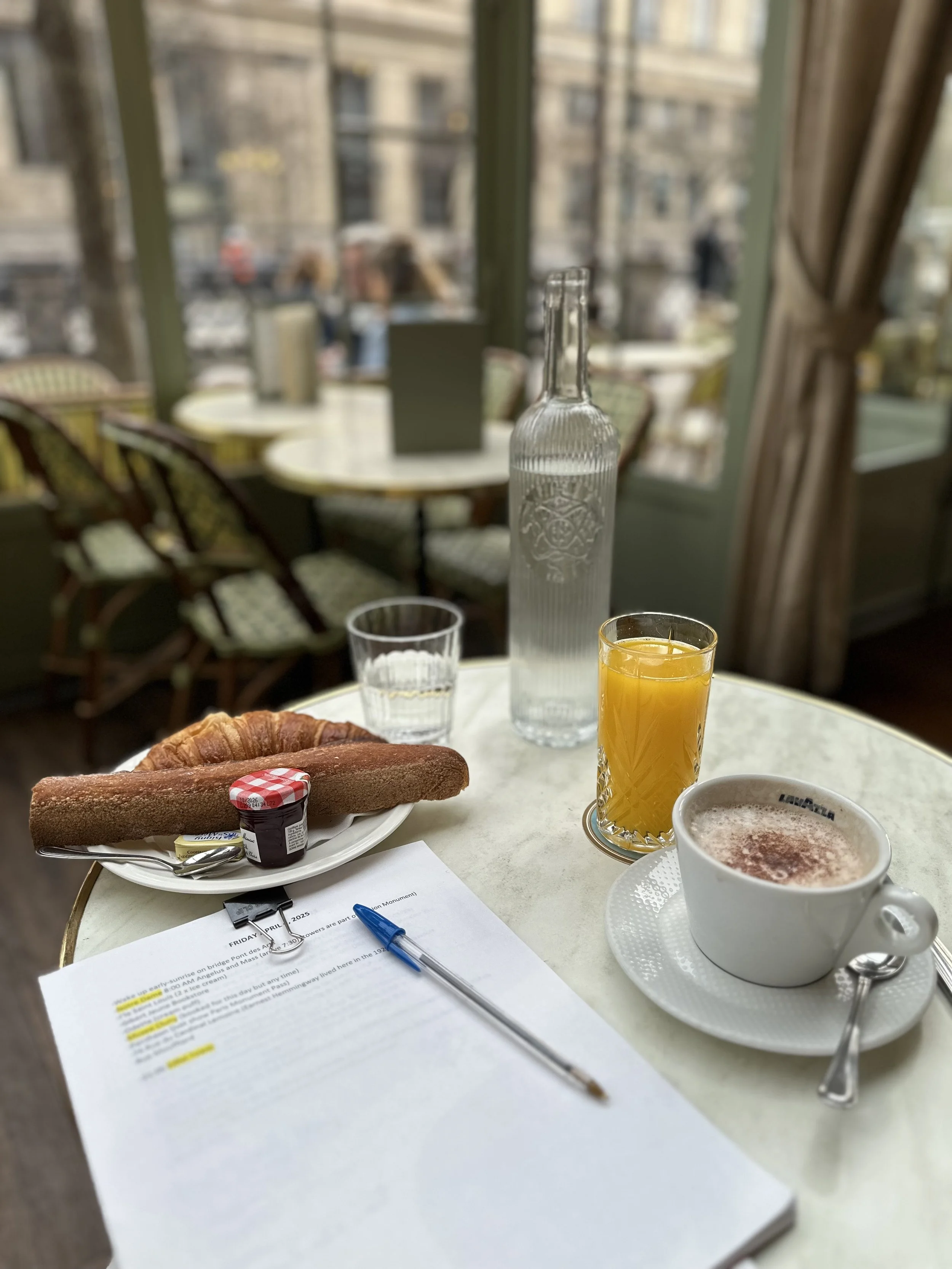 A breakfast table with a croissant, a piece of toast with jam, a glass of orange juice, a cup of coffee, a bottle of water, and a glass of water. There are papers and a pen on the table. The background shows a window with a view of buildings and outdoor scene.