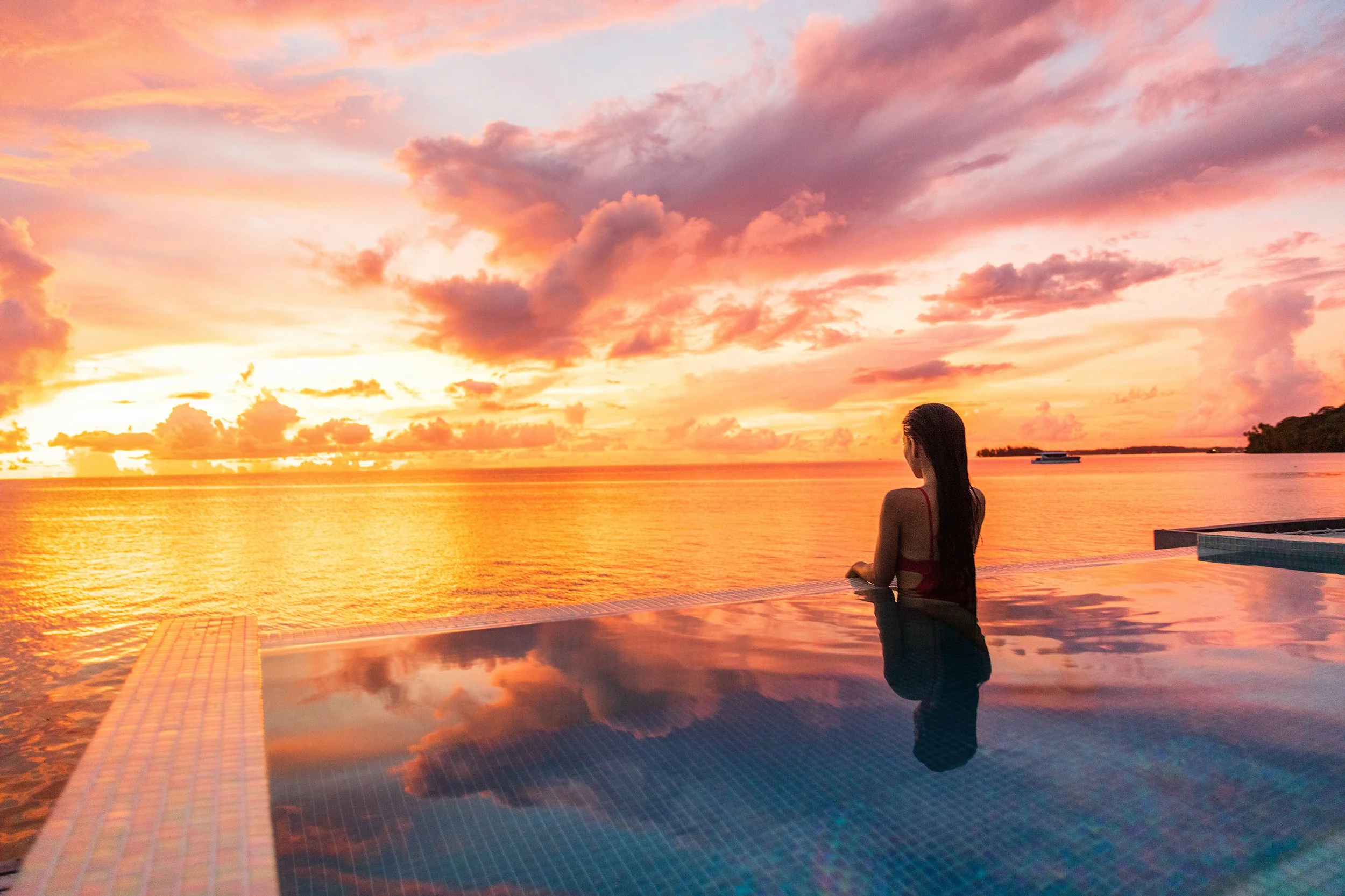 A woman with long dark hair looking out over a sunset by the water, near a swimming pool.