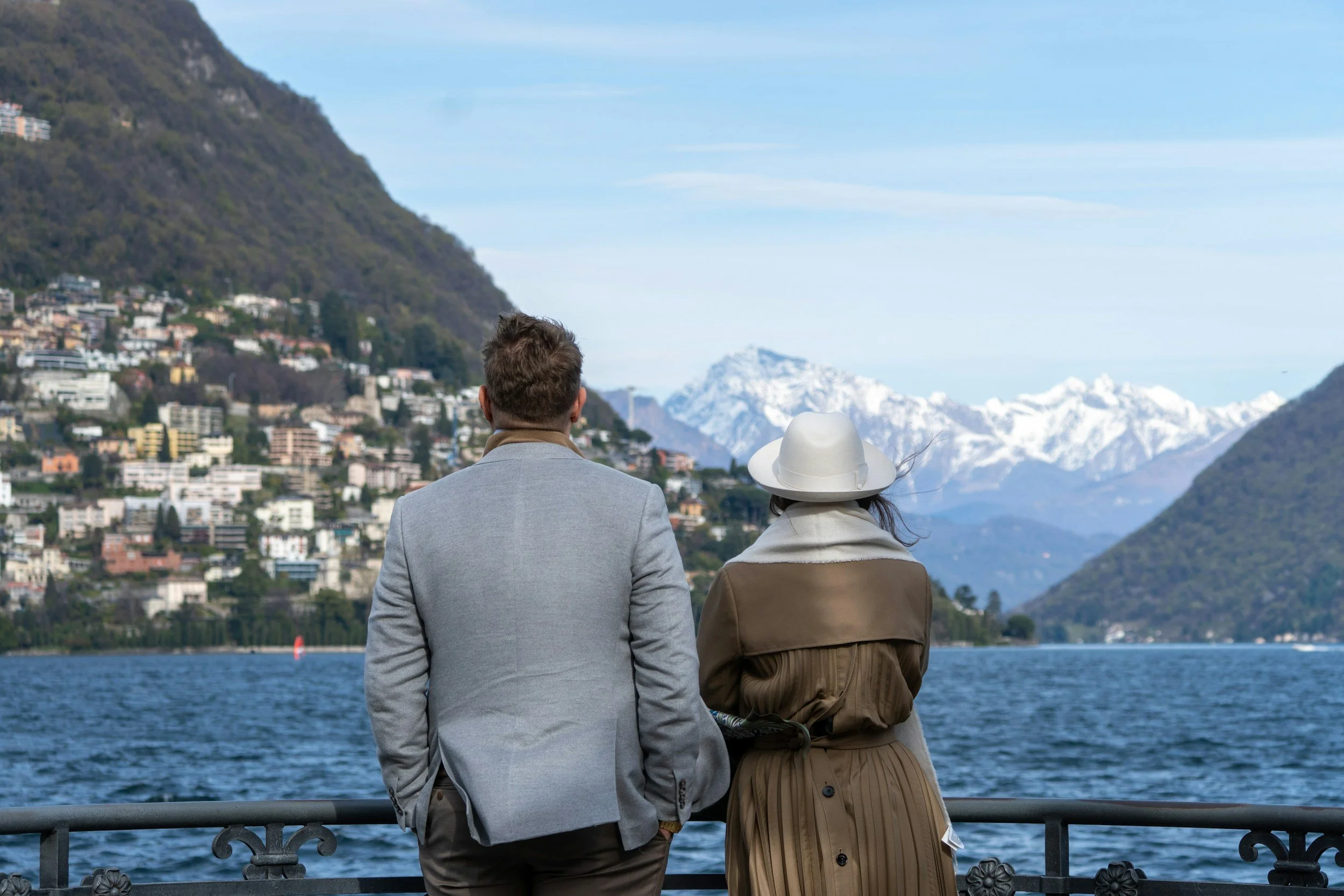 A man and a woman stand by a railing, looking at a lake and mountains in the distance. The man is wearing a light gray jacket, and the woman is wearing a beige coat and a wide-brimmed white hat.