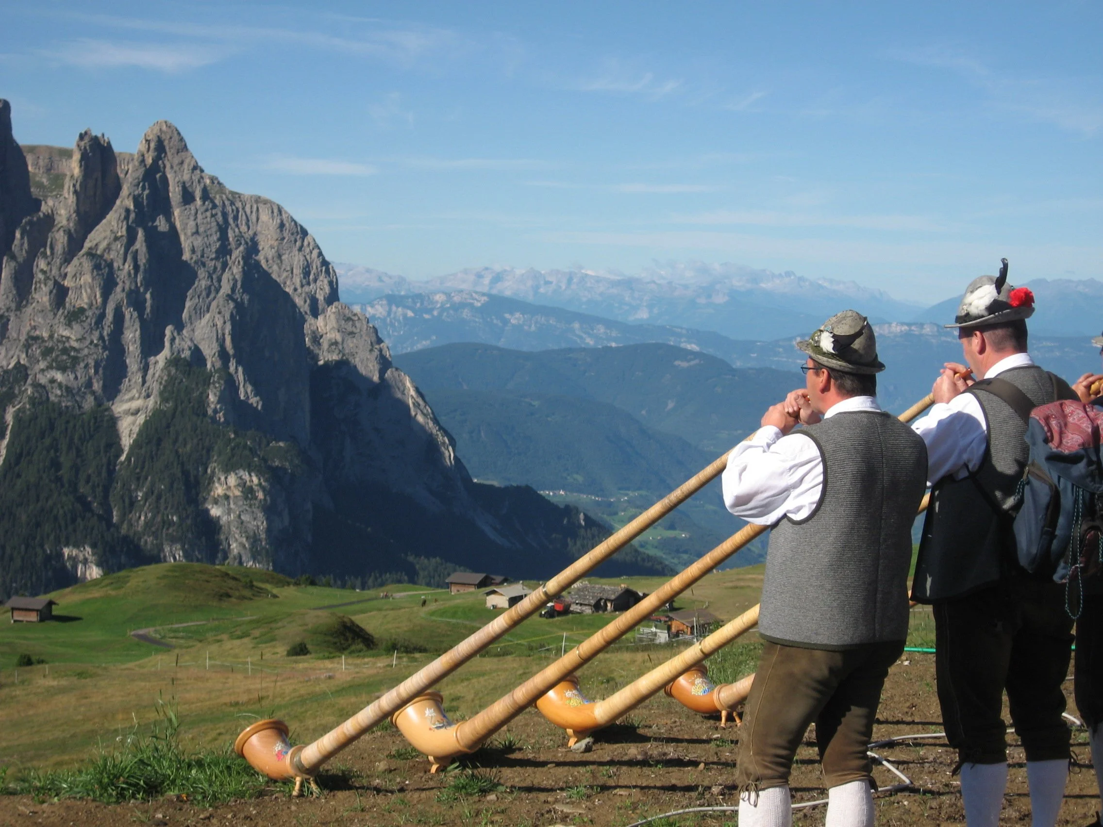 Group of men wearing traditional Bavarian clothing, playing alphorns outdoors in a mountainous landscape with green fields and rugged peaks in the background.