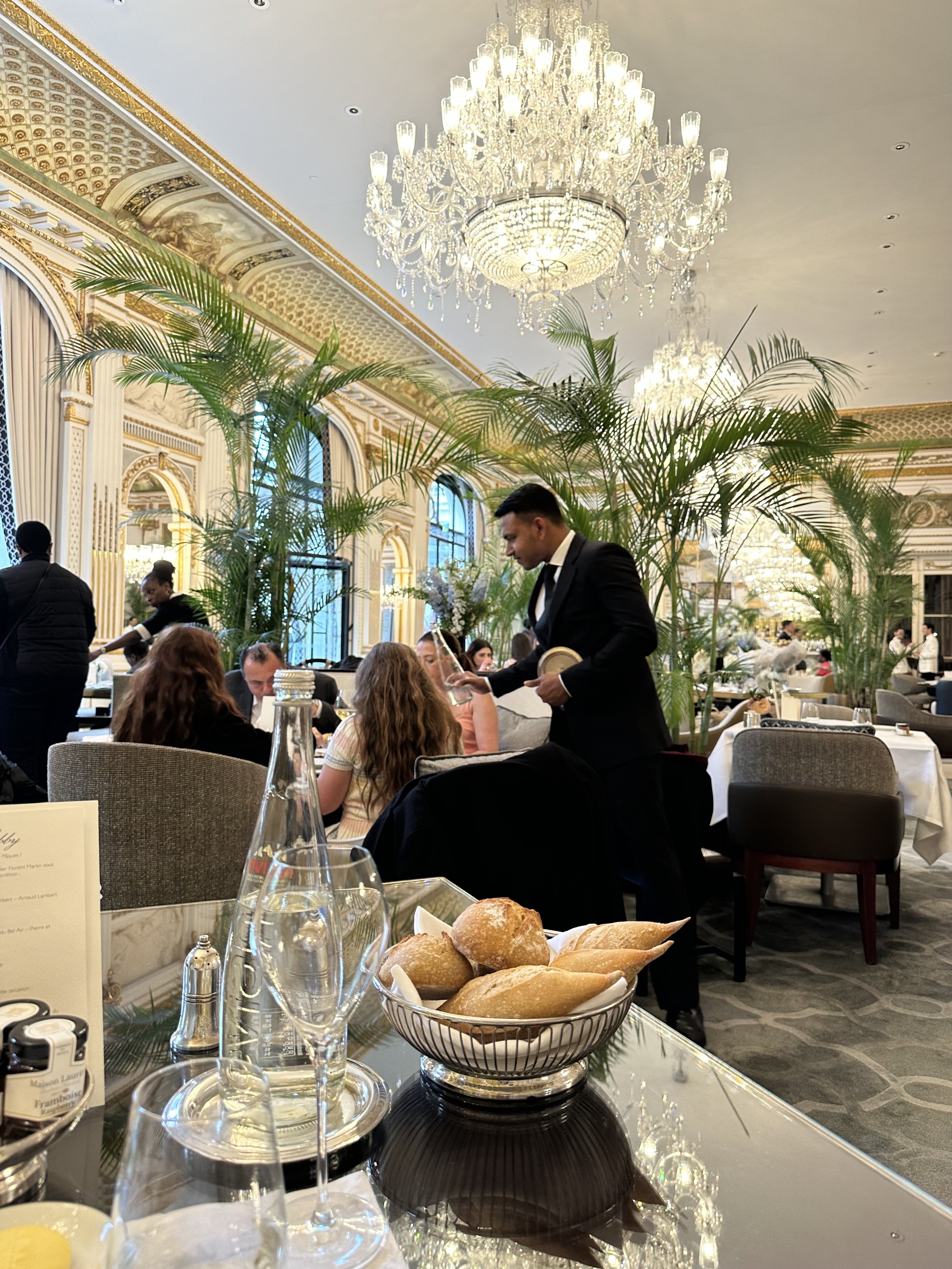 Interior of an elegant restaurant with a crystal chandelier, large windows, green plants, and people dining, with bread baskets and glasses on the table in the foreground.
