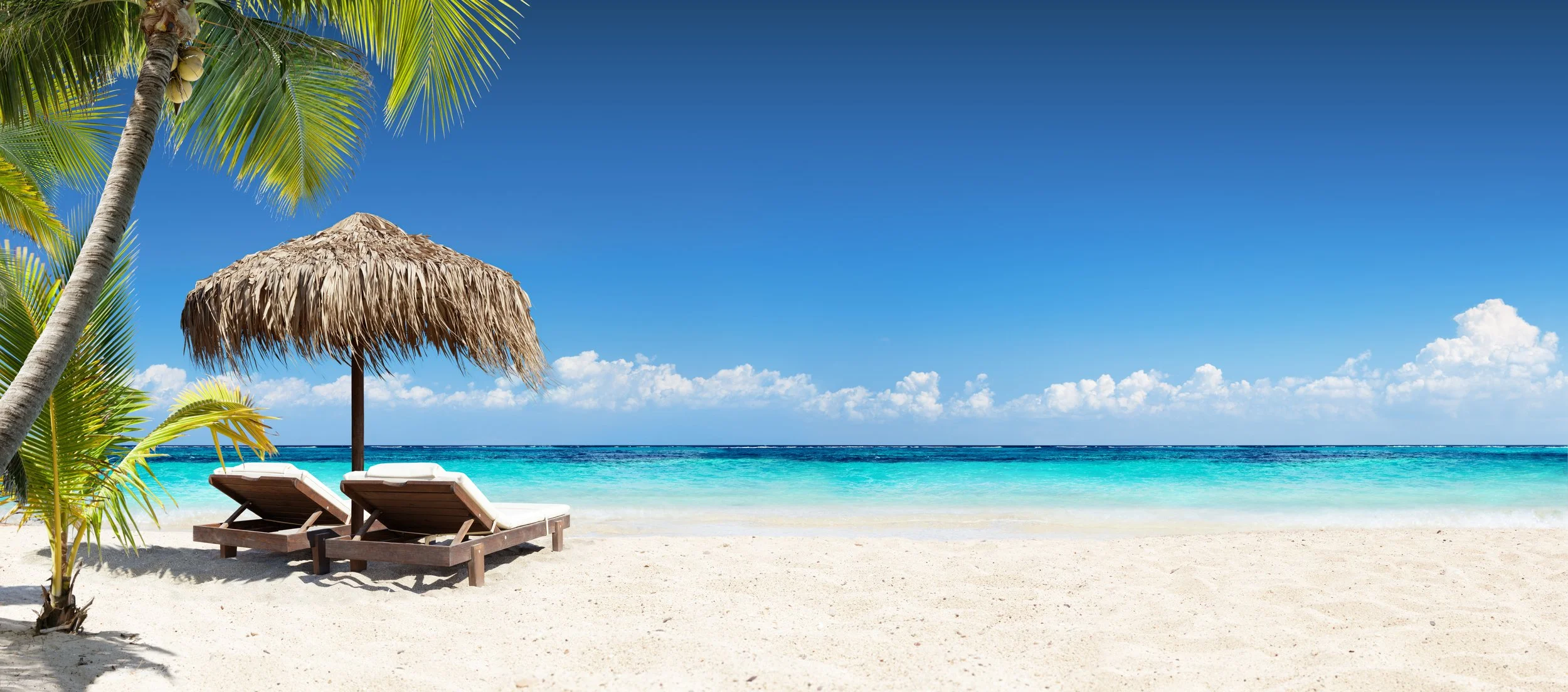 A tropical beach scene with white sand, turquoise water, a thatched umbrella, and two lounge chairs, with palm trees and a blue sky with white clouds.
