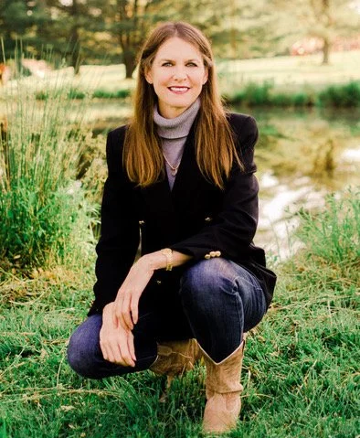A woman with long brown hair, wearing a black blazer, gray turtleneck, and jeans, crouching outdoors near a pond with trees and greenery in the background.