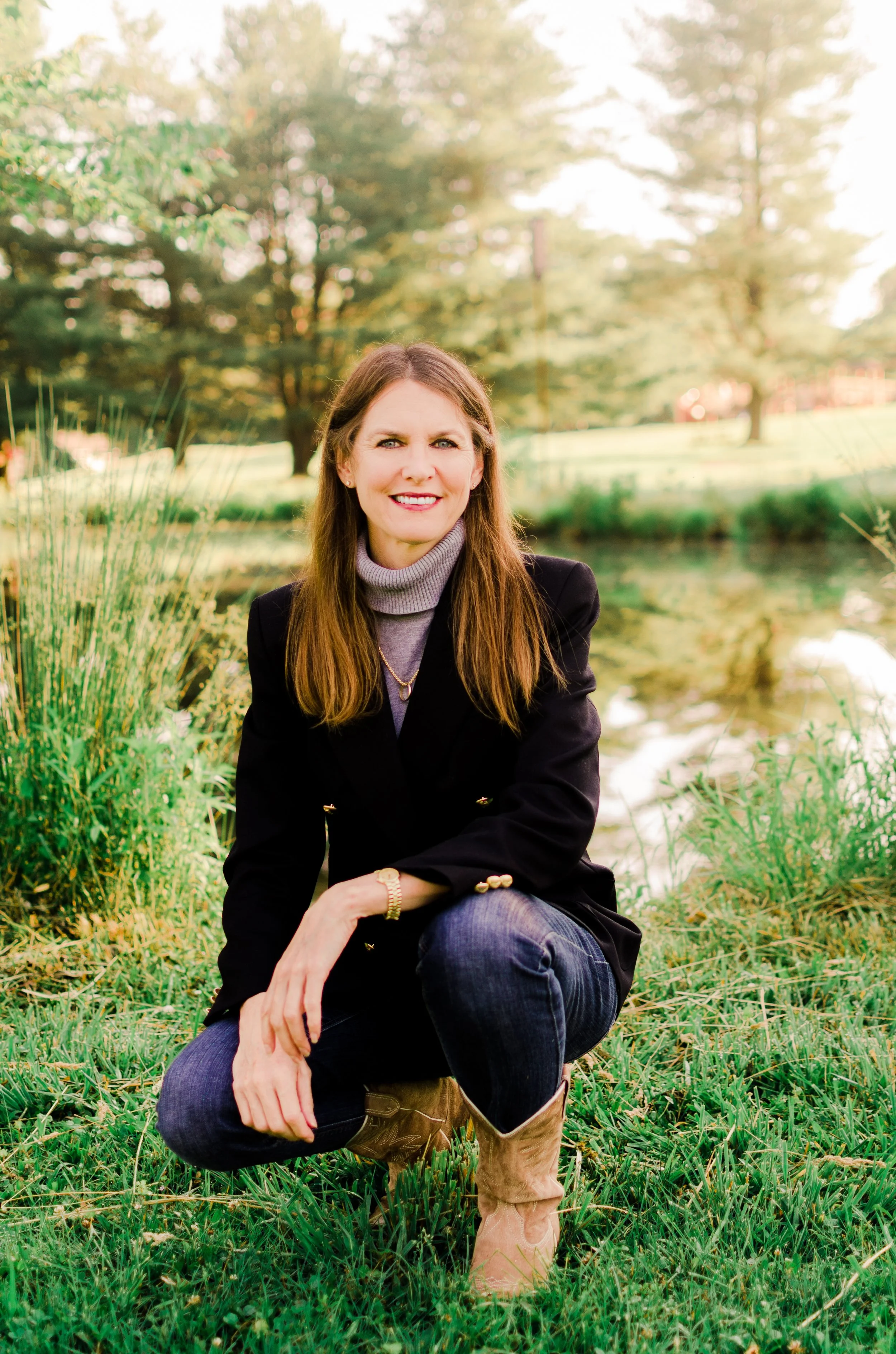 A woman with long brown hair sitting outdoors near a pond in a grassy area, smiling at the camera, wearing a black blazer, gray turtleneck, blue jeans, and cowboy boots, with trees in the background and sunlight filtering through.