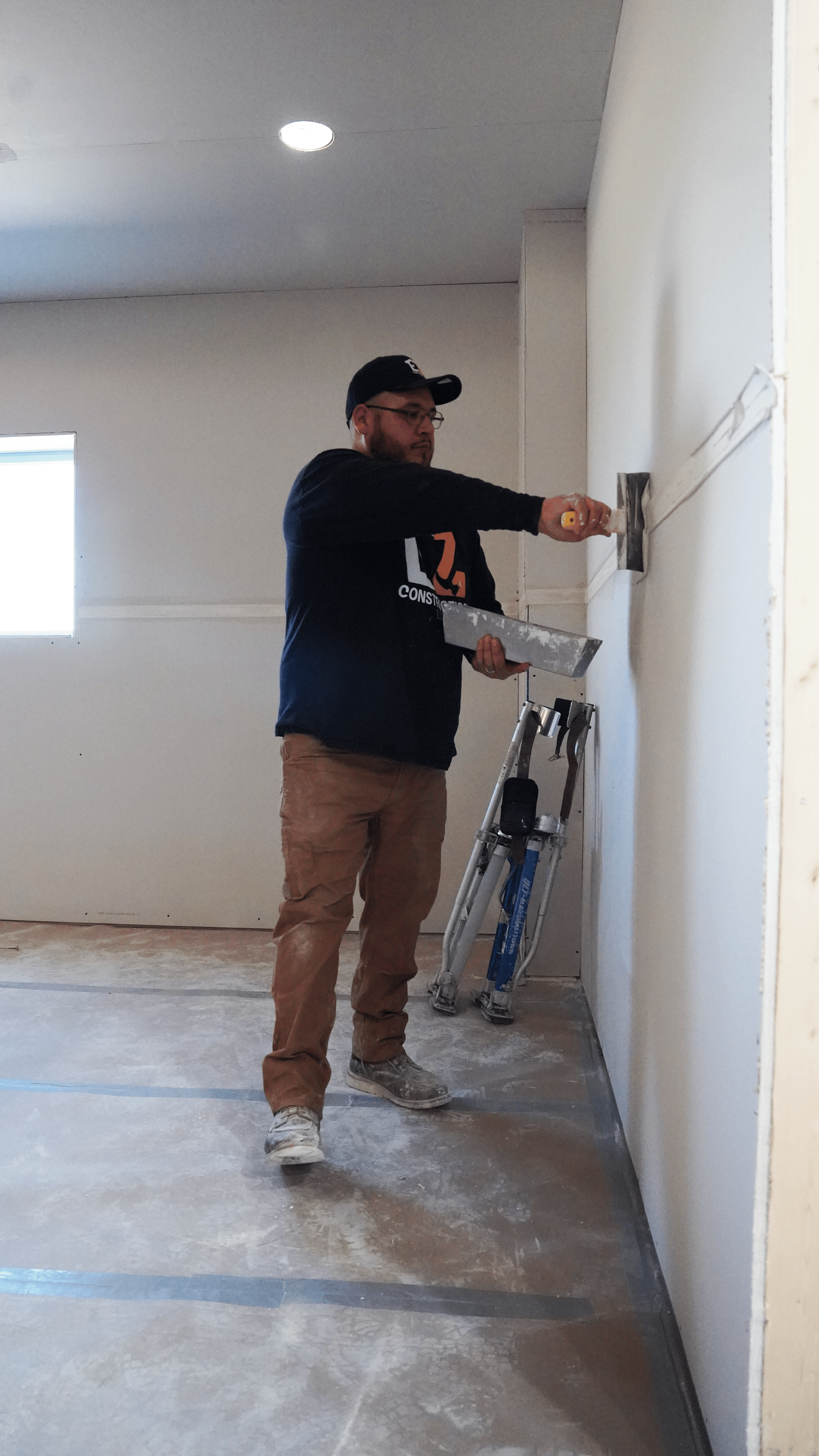 A man in a black hoodie and brown pants using a putty knife to scrape drywall in a room under construction, with a stepladder and a small window in the background.