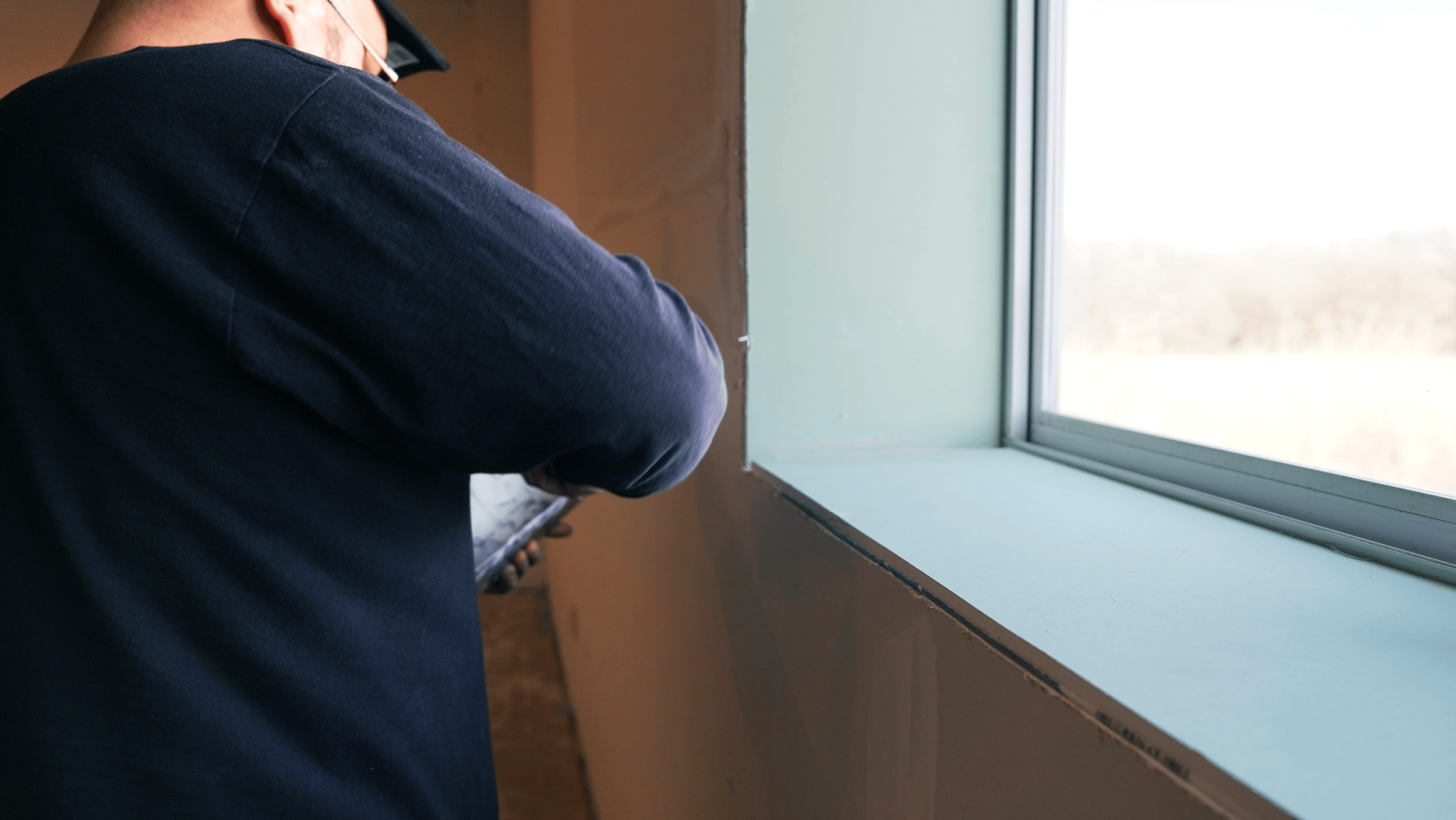 Person wearing a dark blue shirt, working near a window with a view outside, holding a tool for drywall..