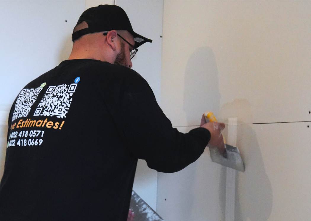A man wearing a black cap, glasses, and a black long sleeve shirt is applying joint compound to a wall with a putty knife.