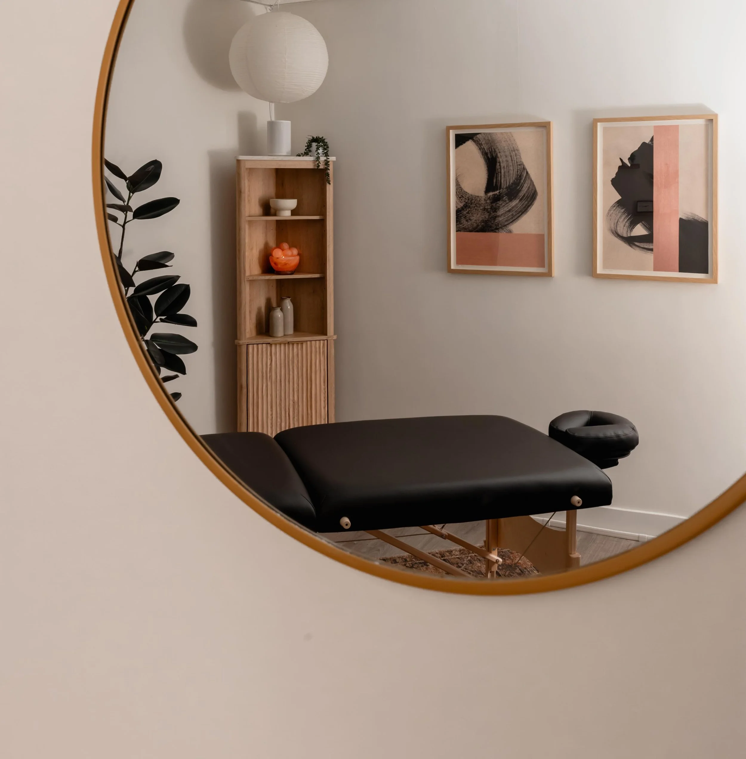Reflection of Nereya's massage room with a black massage table, wooden shelving with decorative objects, framed abstract art, and a hanging paper lantern, seen through a round mirror.