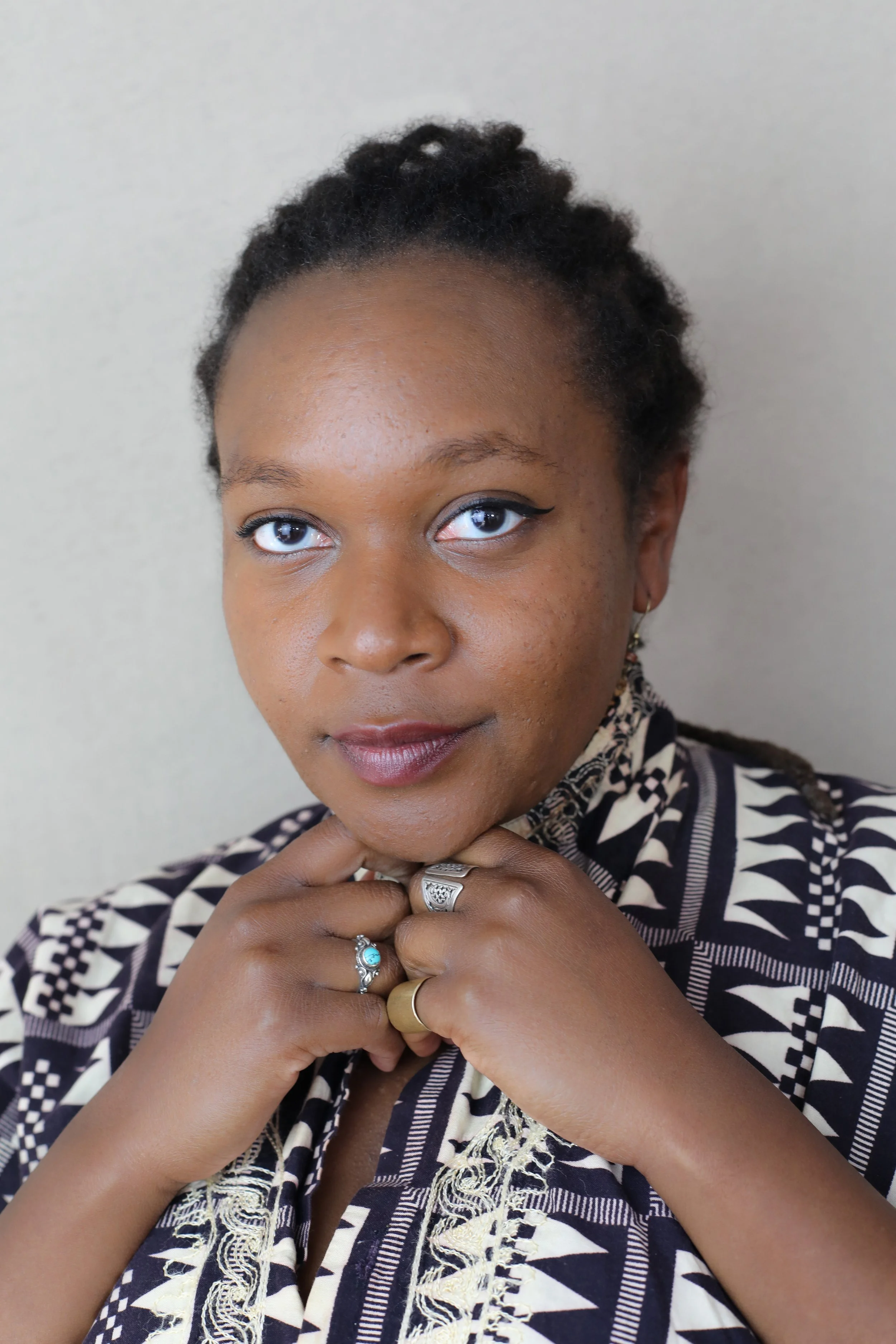 Close-up portrait of Nereya: a dark-skinned woman with light makeup, hair pulled back, dressed in a patterned black and white outfit, posed with her hands near her chin, standing against a plain background.
