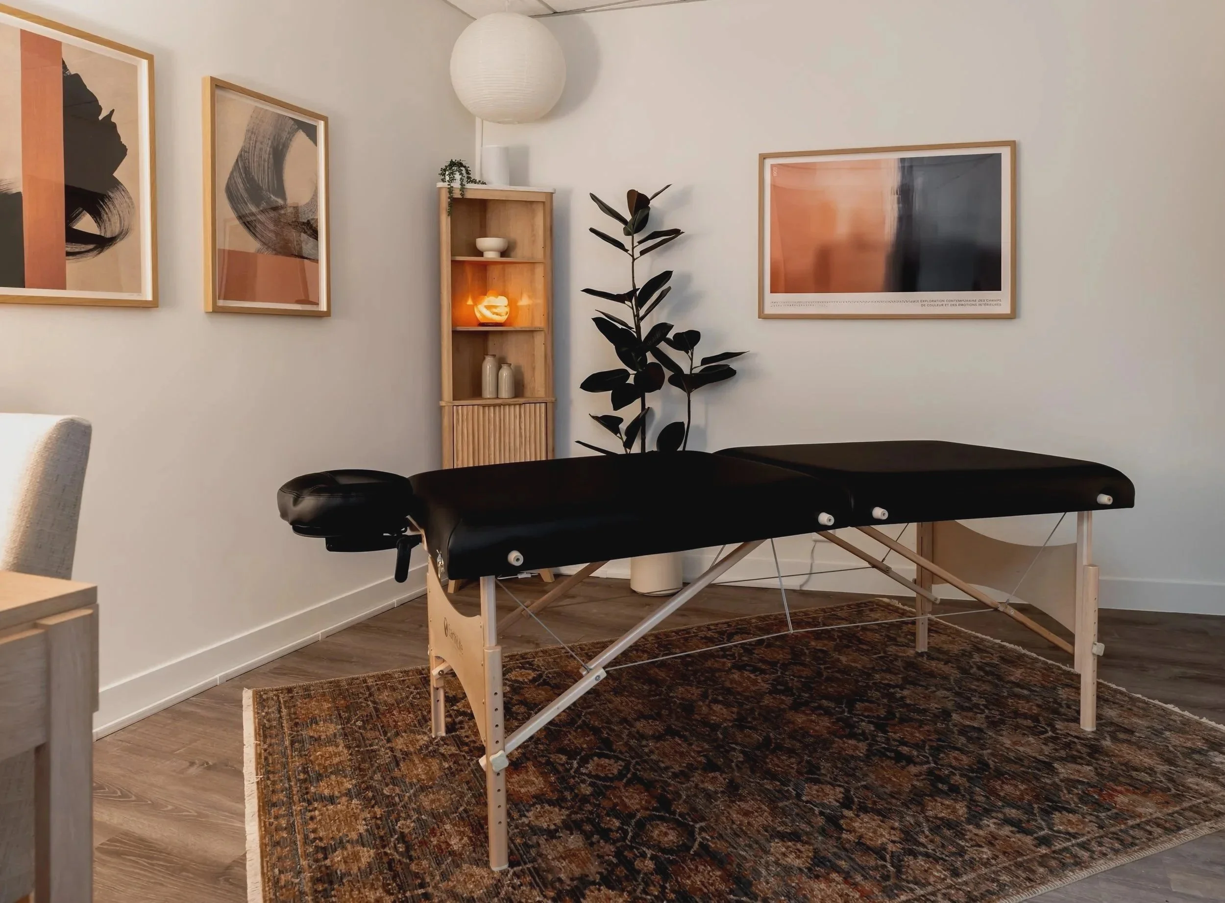 An image of Nereya's workspace with a massage table in a modern room with framed abstract art on the walls, a small wooden shelving unit, a large potted plant, and a patterned area rug.