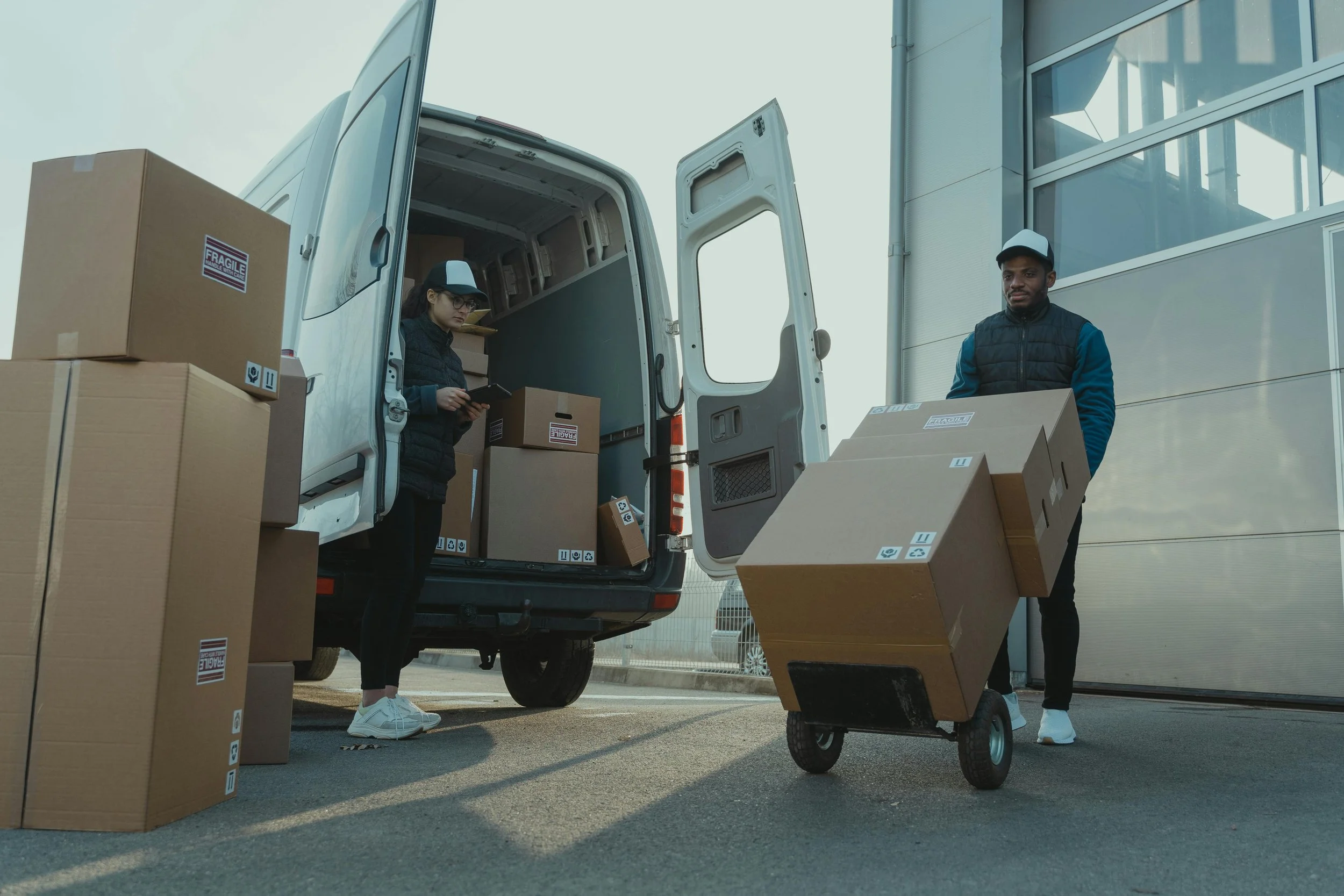 Two delivery workers unloading boxes from a white delivery van outside a building, with one person standing by the van's open door and the other pushing a hand truck loaded with boxes.