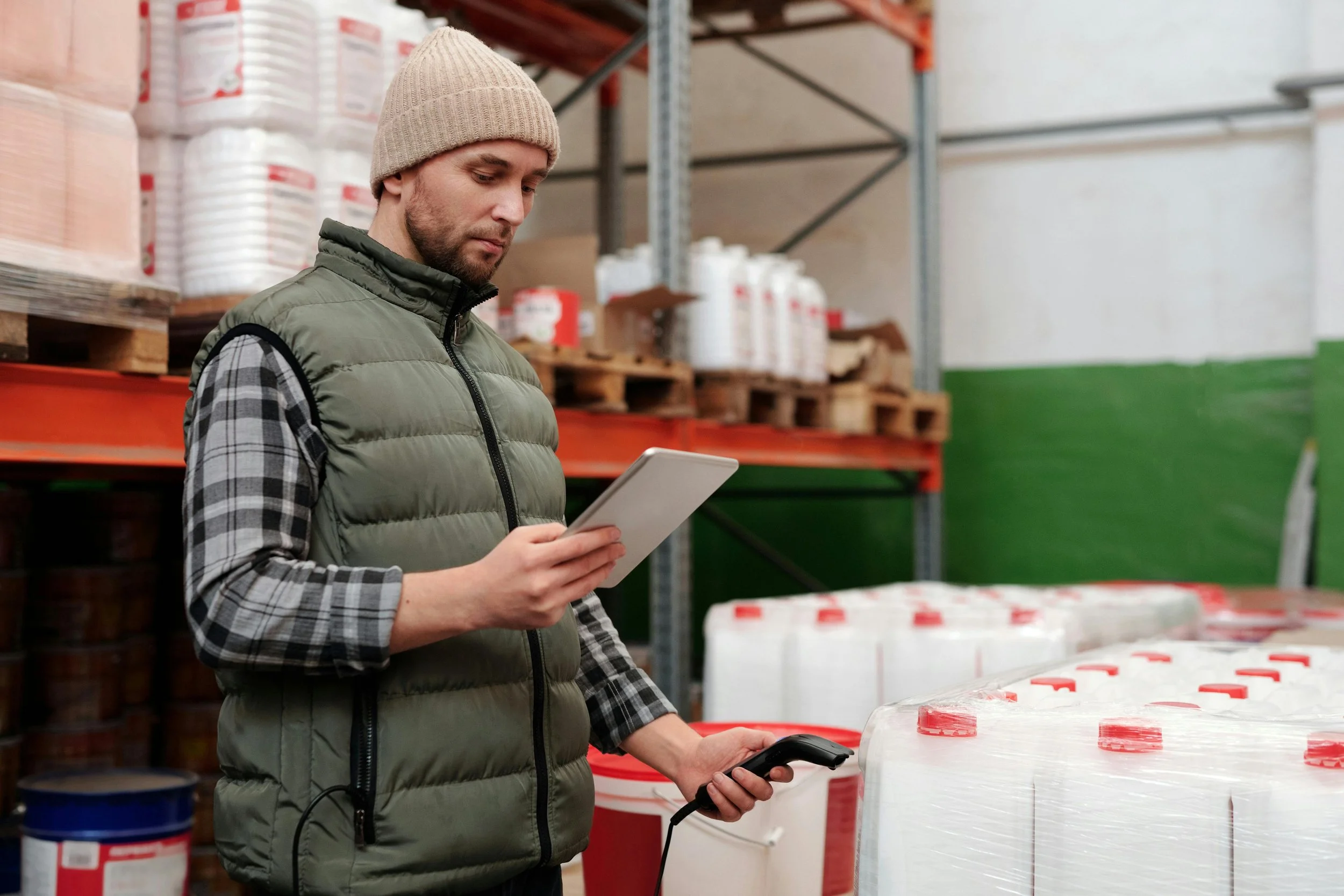A man in a beige beanie, green puffer vest, and plaid shirt holding a tablet and a barcode scanner in a warehouse aisle with shelves of large white containers and pallets.