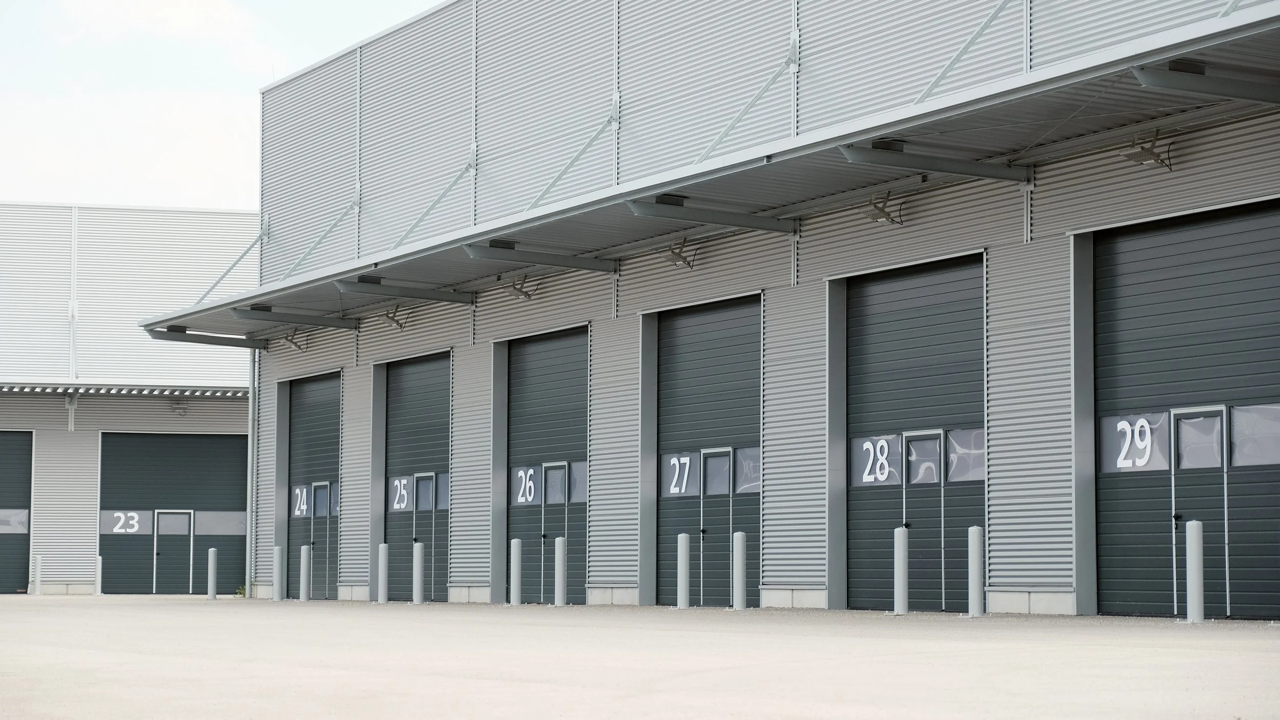 Industrial storage units with closed roll-up doors numbered 23 to 29, metal exterior, and safety bollards in front.