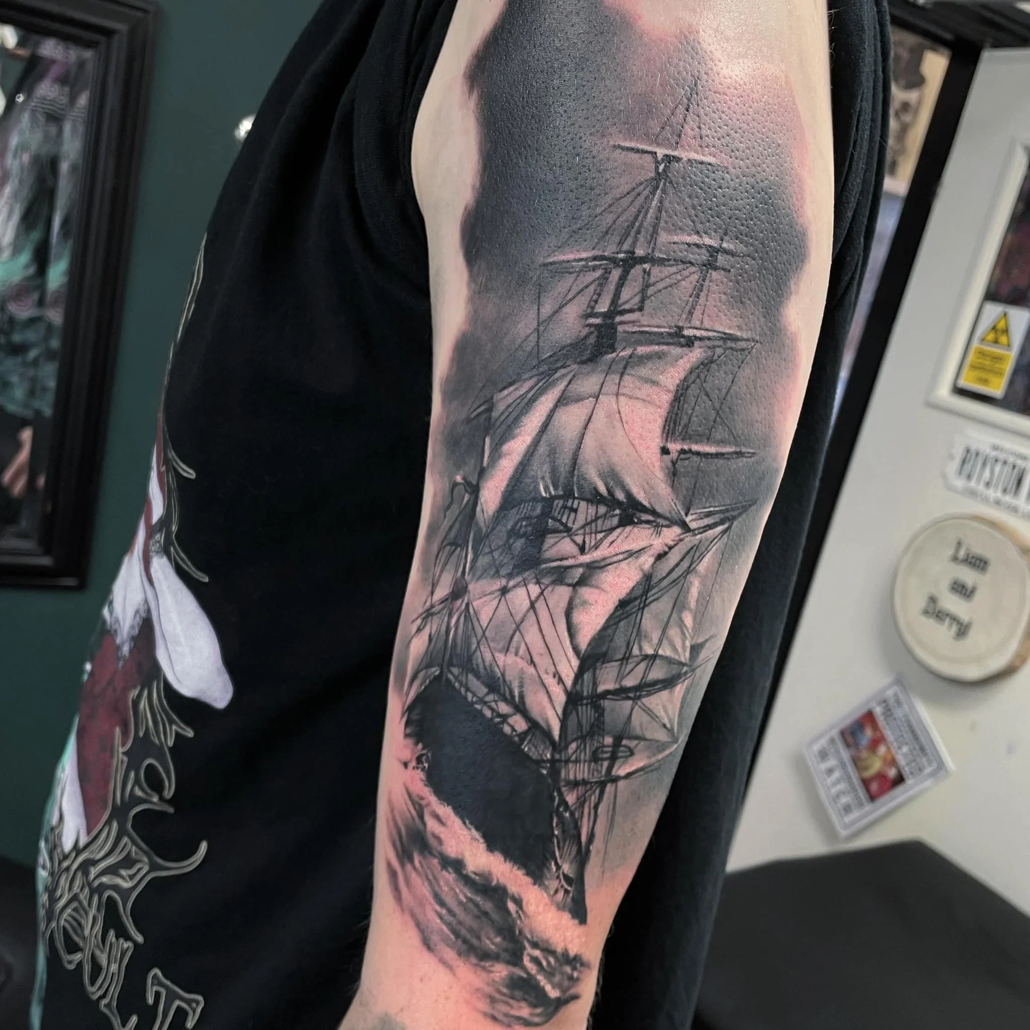 Tattoo of a sailing ship on person's upper arm, depicted with detailed sails, rigging, and a realistic sea environment at the bottom.