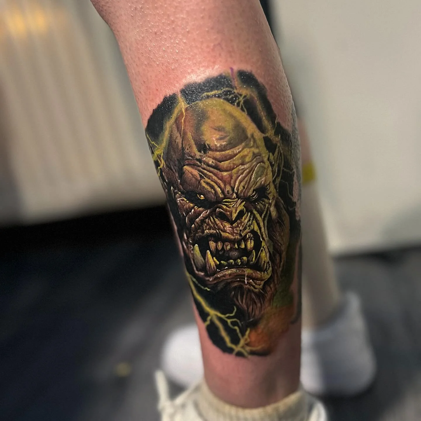 Tattoo of an angry, monstrous face, resembling a giant gorilla or orc, with fierce expression and sharp teeth, surrounded by yellow lightning bolts.