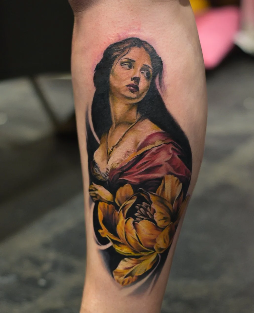 A colored tattoo of a woman with dark hair, wearing a necklace and draped in a red and yellow cloth, with a large yellow flower below her.