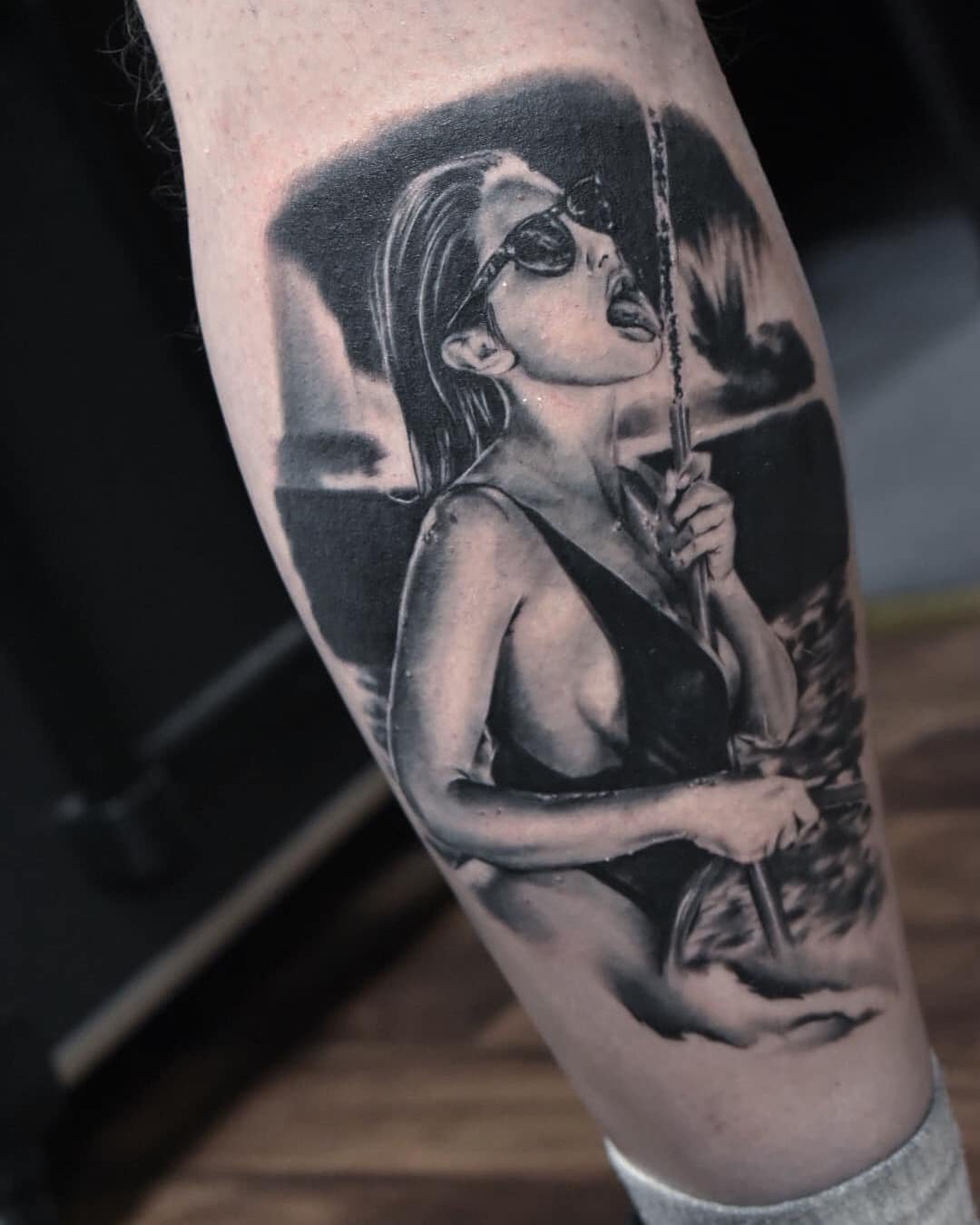 A detailed black and grey tattoo of a woman with sunglasses and her tongue out, holding an umbrella, wearing a low-cut top, on a person's arm.