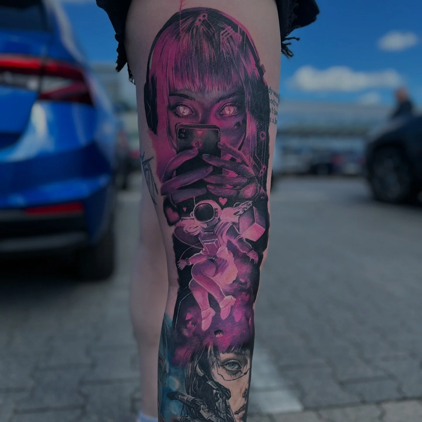 Tattoo of a woman with pink hair and headphones taking a selfie, with a small astronaut figure below, depicted in shades of purple, pink, and black.