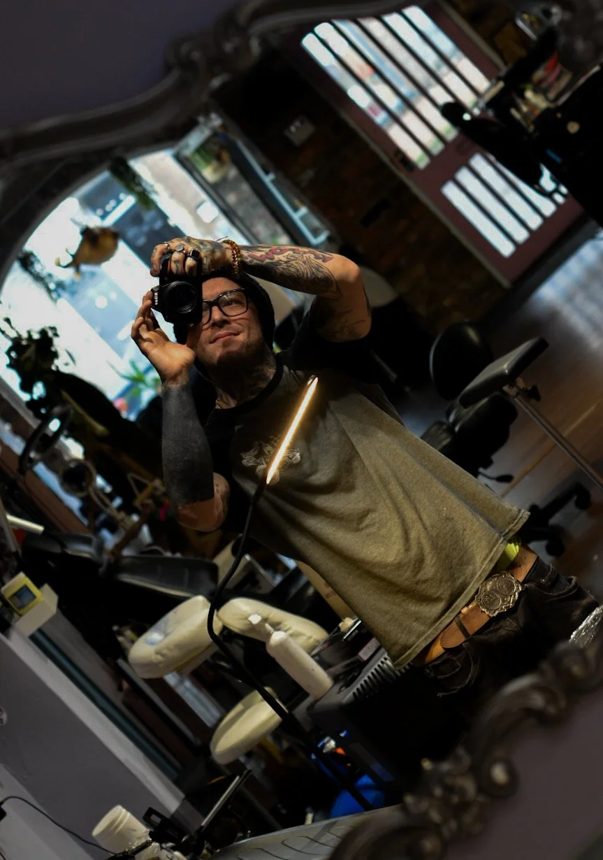 A man with tattoos and glasses taking a selfie with a camera in a mirror inside a tattoo studio.