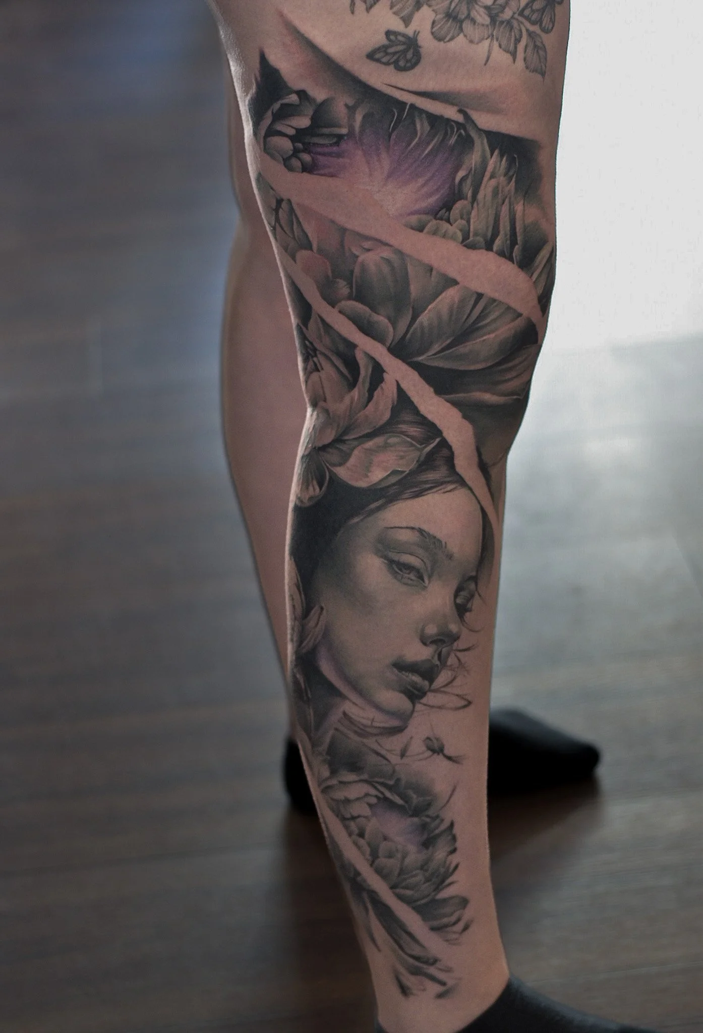 Tattooed leg with a detailed black and gray portrait of a woman and floral elements, with a torn paper effect over the image.