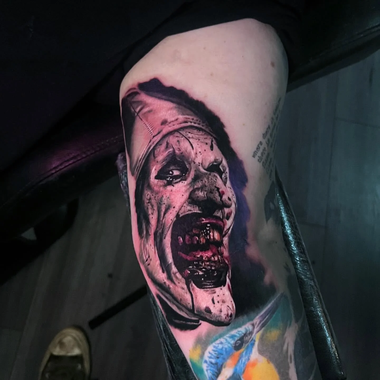 Tattoo of a disturbing clown face with yellow teeth, dark eyes, and an open mouth showing teeth, on person's leg.