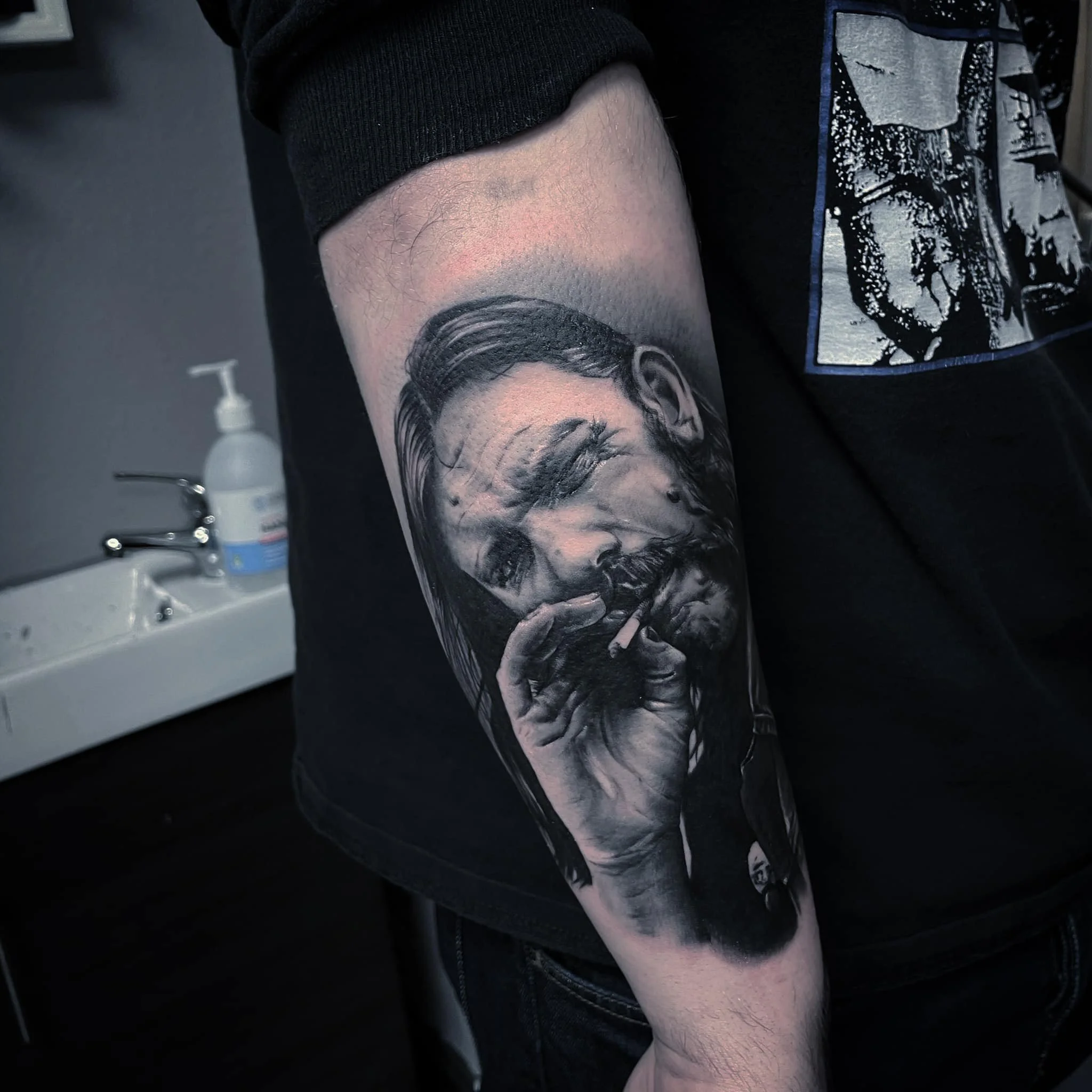 Black and grey realism tattoo of a man with long hair and a beard, smoking a cigarette, on a person's arm.