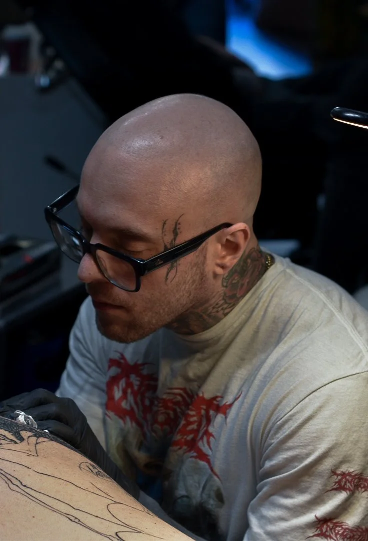 A male tattoo artist with a shaved head and glasses working on a tattoo on a client's arm.