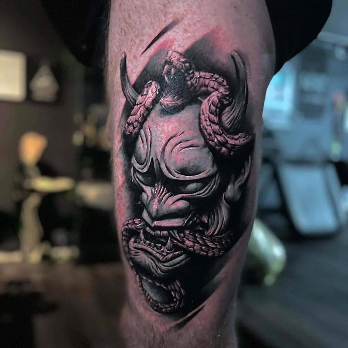 Black and gray tattoo of a demonic face with horns, an angry expression, and a snake wrapped around its nose and mouth.
