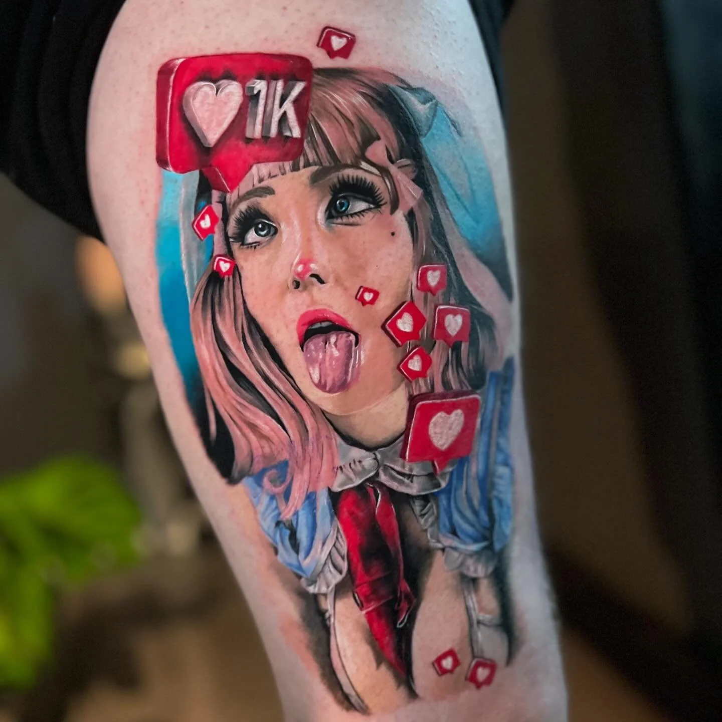 Tattoo of a woman with pink hair sticking out her tongue, surrounded by red social media notification icons with hearts and a "1K" symbol, on a person's arm.