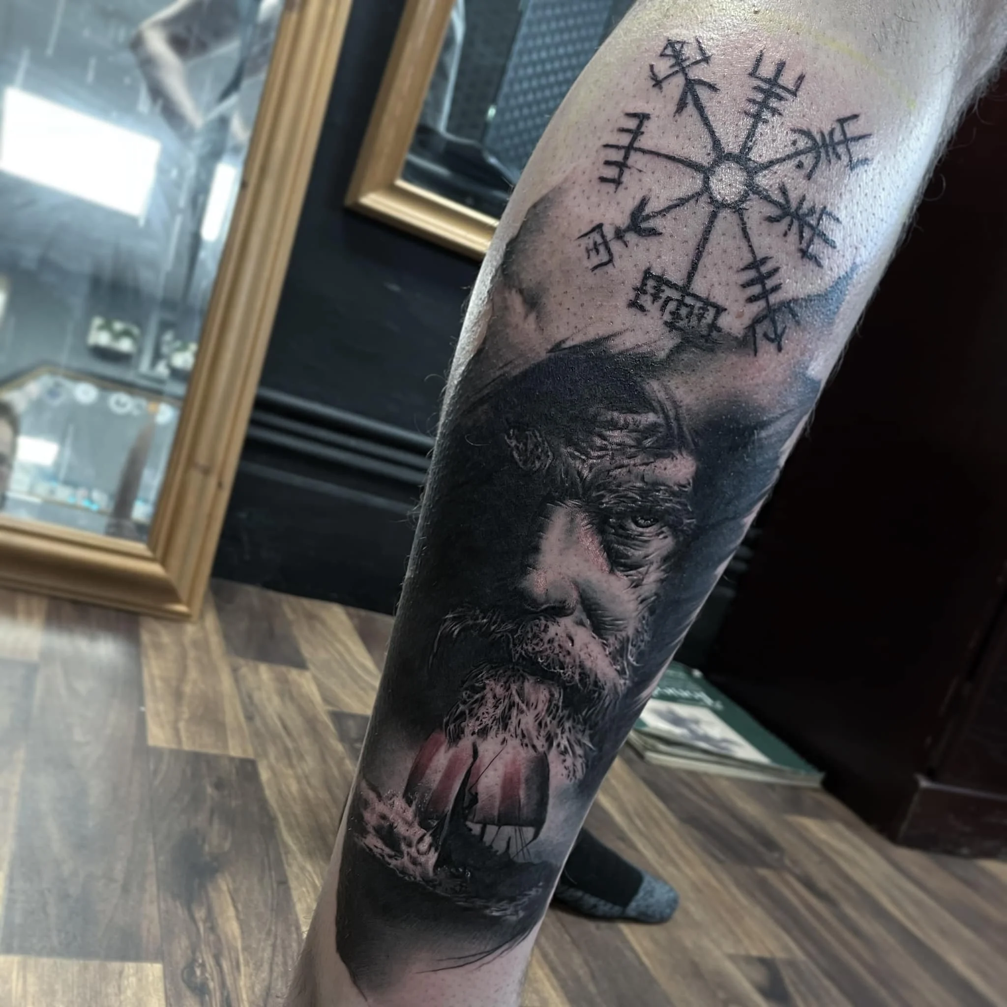 Tattoo of a bearded man's face with a ship sailing on turbulent waters underneath, and a Viking-style compass or symbol with runes or symbols above.