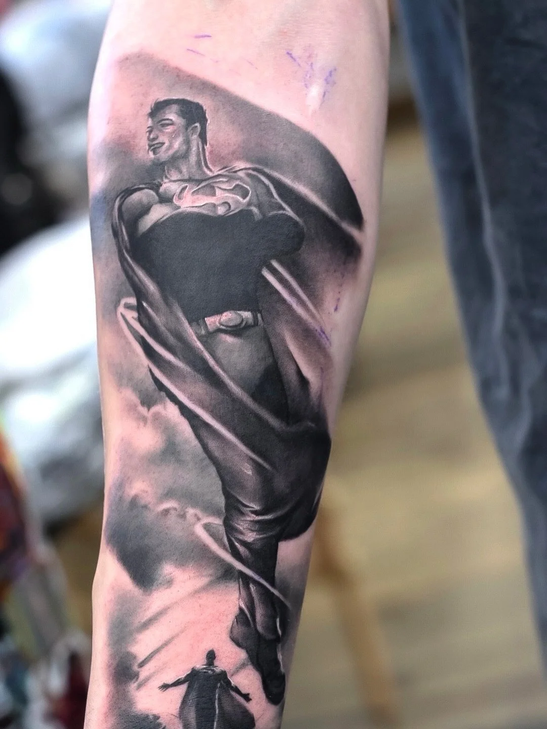 Black and gray tattoo of Superman with a smiling face, iconic costume, and the Superman logo on the chest, flying with a swirl background.